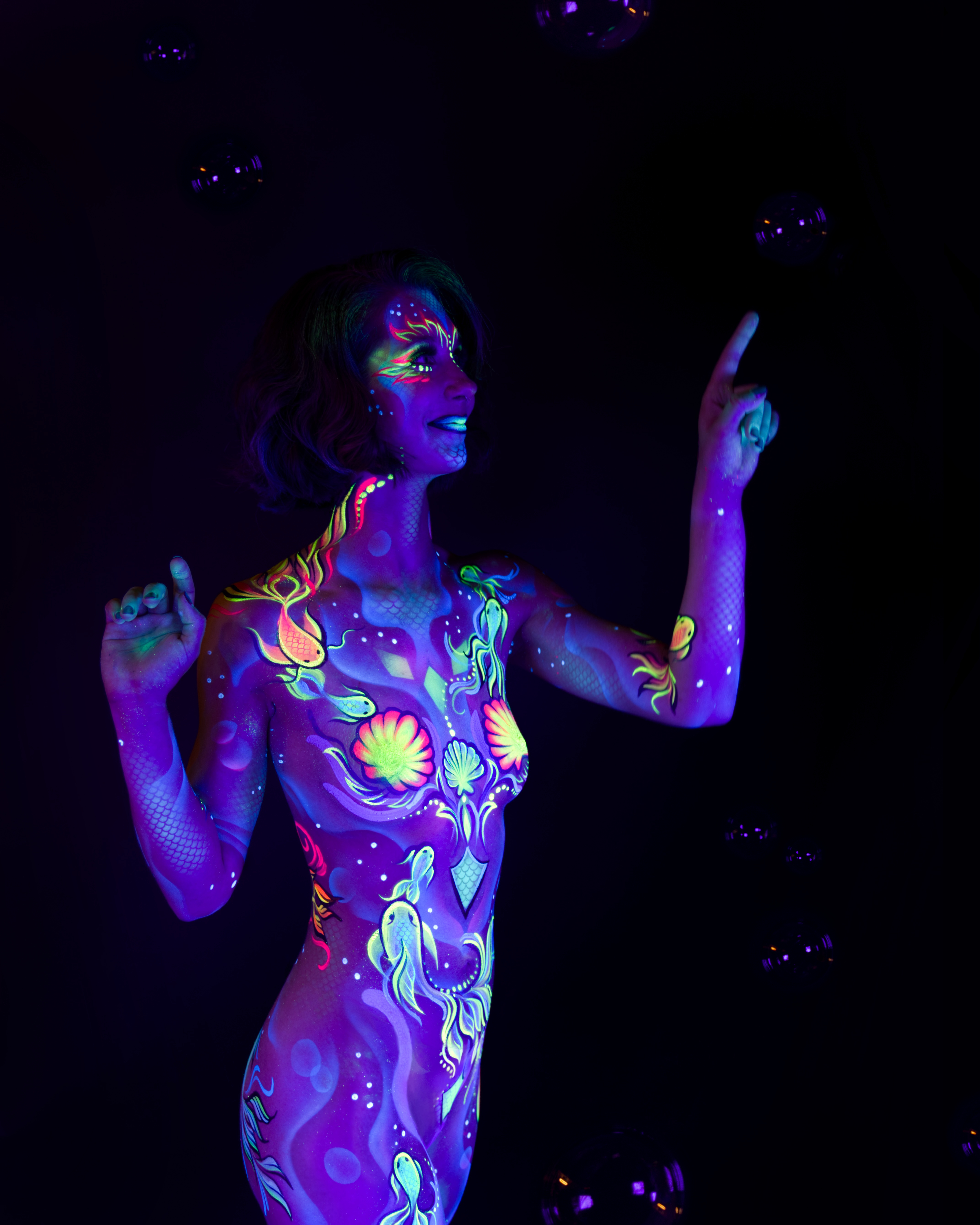 Karoline stands in a three-quarter pose with bubbles visible in the background. The bodypaint depicts an underwater sealife theme, including shell and fish motifs rendered in pink, green and blue across the torso and legs. Bodypaint by Joana Rohr. Photographed by Elad Itzkin Photography under UV light in his studio in Gesundbrunnen, Berlin.