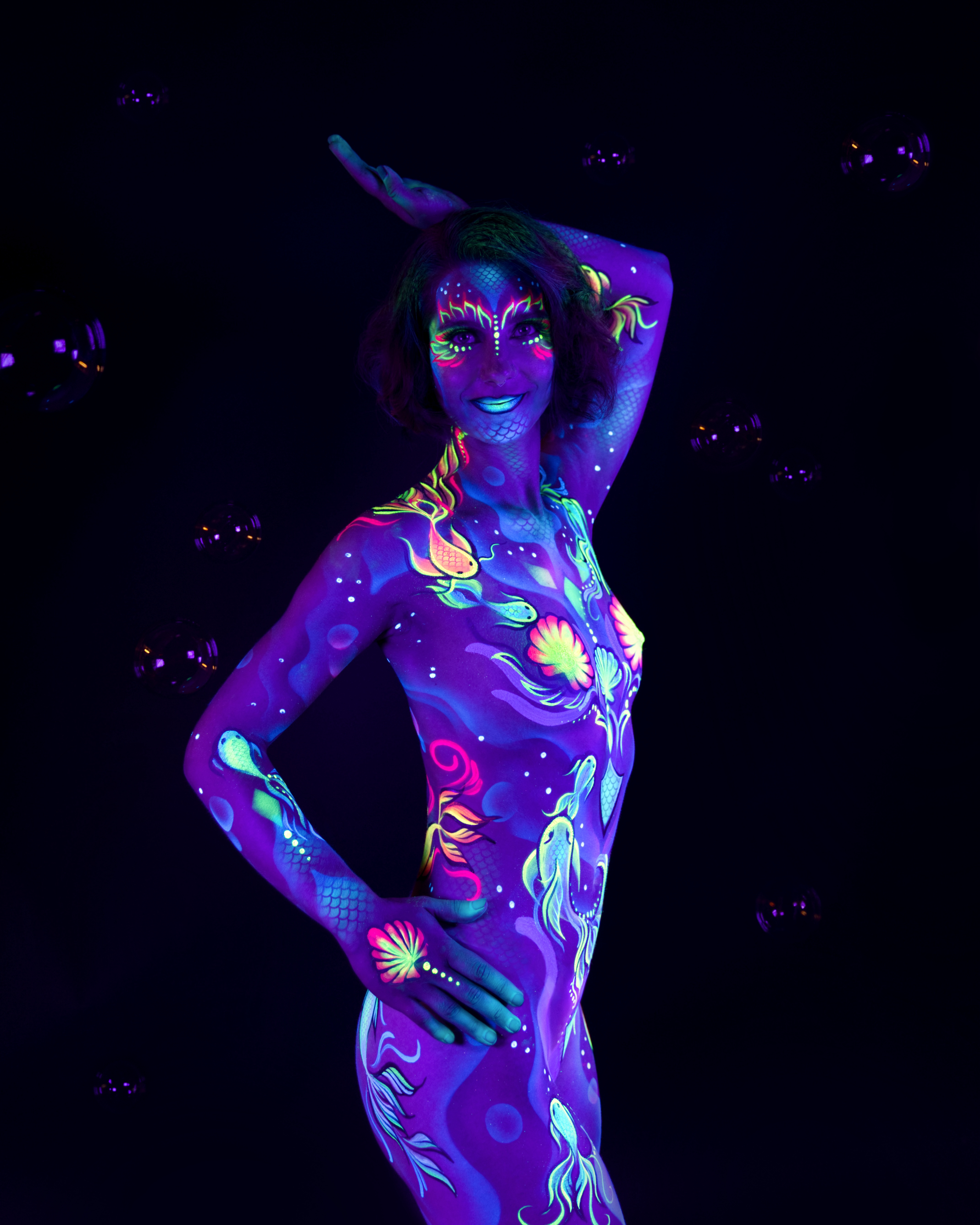 Karoline stands in a three-quarter pose with the head turned upward and to the side, bubbles scattered around her. The sealife-themed bodypaint combines shell, fish and coral motifs in pink, green and blue across the body. Bodypaint by Joana Rohr. Photographed by Elad Itzkin Photography under UV light in his studio in Gesundbrunnen, Berlin.