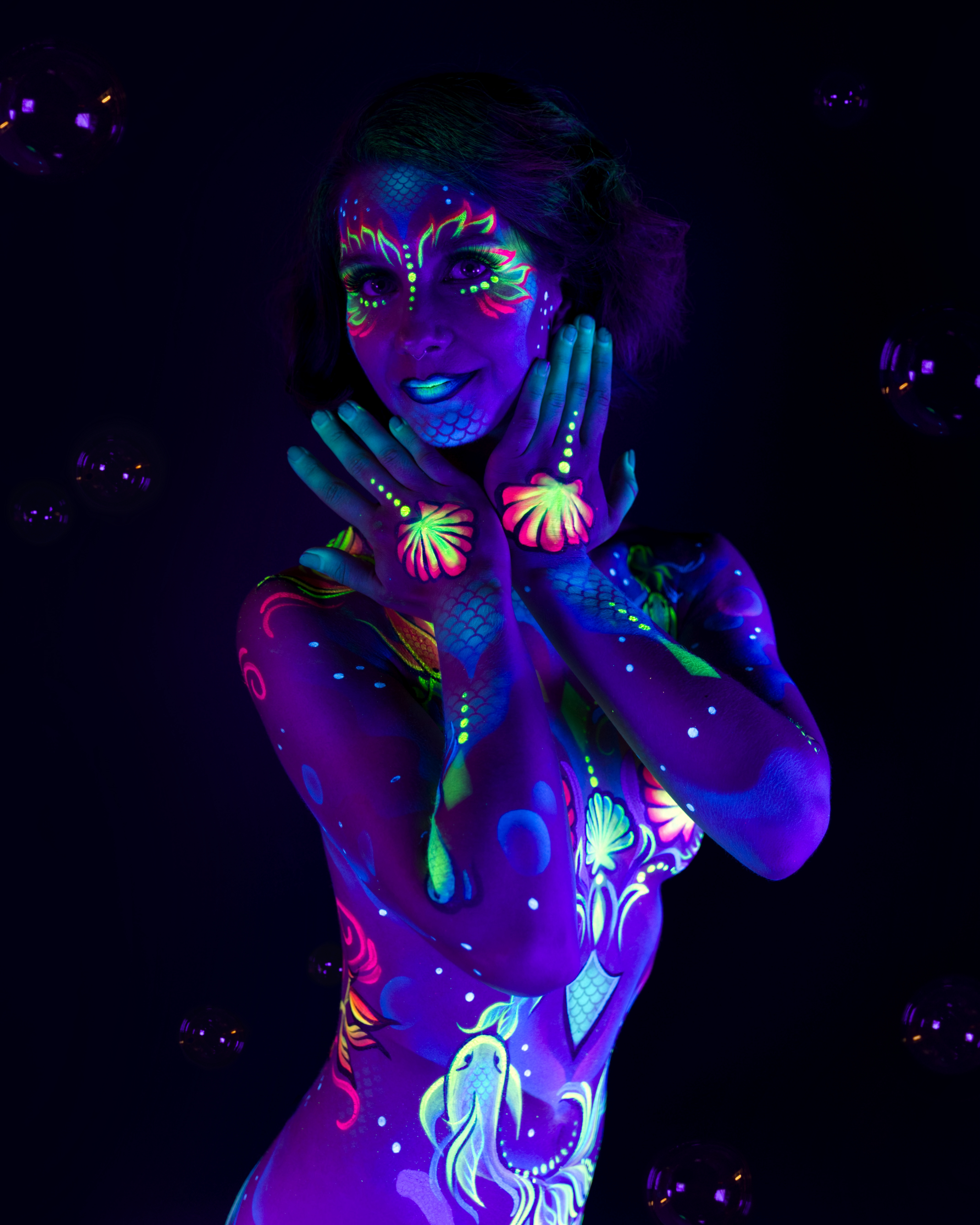 Karoline is shown in a close upper-body crop, facing the camera. The sealife-themed bodypaint includes shell and fish motifs in pink, green and blue, with a scale-like pattern extending across the neck and chest. Bodypaint by Joana Rohr. Photographed by Elad Itzkin Photography under UV light in his studio in Gesundbrunnen, Berlin.