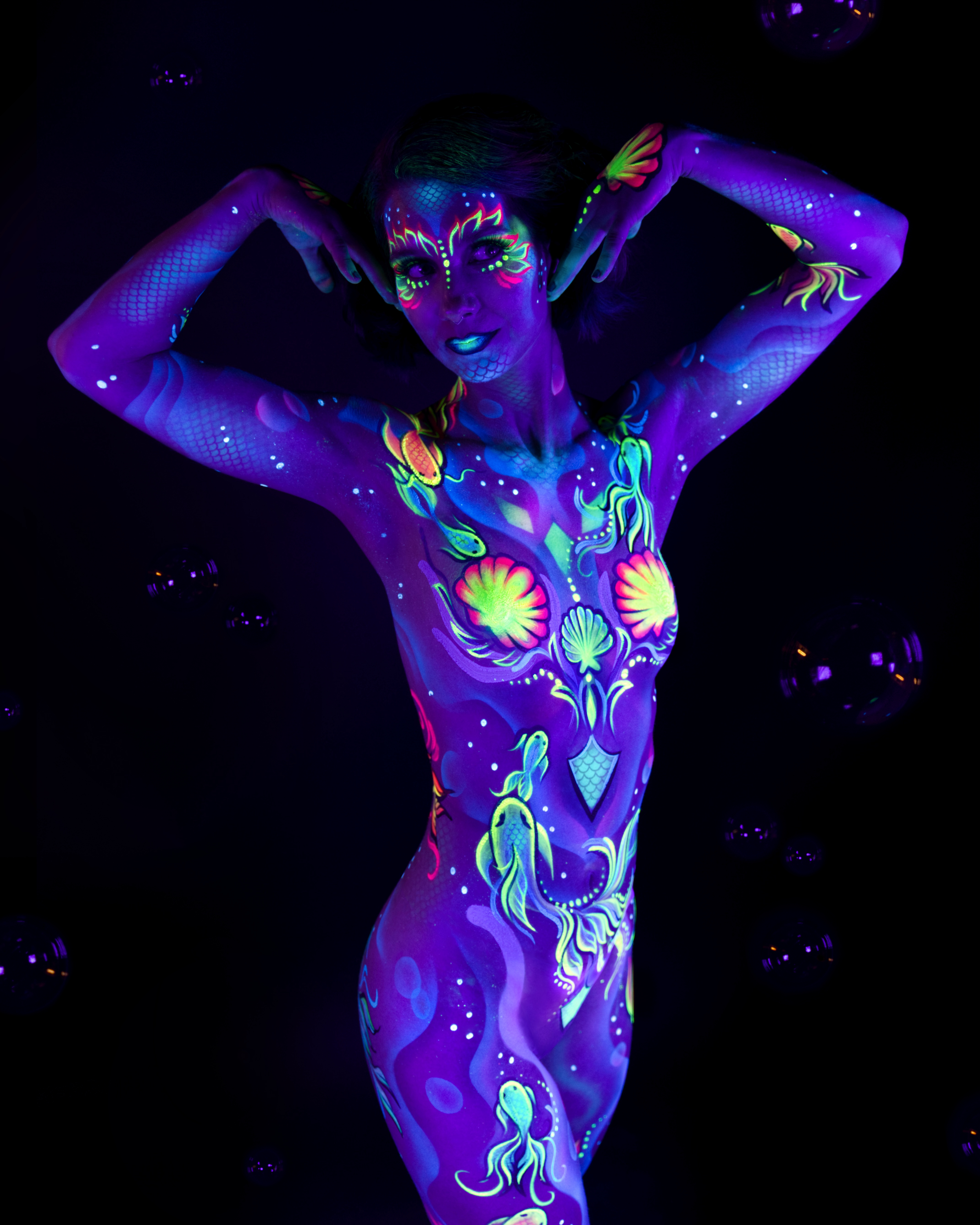 Karoline stands facing the camera in a full-body pose. The bodypaint covers the body in an underwater theme, combining shell, fish and seahorse motifs in pink, green, blue and yellow. Bodypaint by Joana Rohr. Photographed by Elad Itzkin Photography under UV light in his studio in Gesundbrunnen, Berlin.
