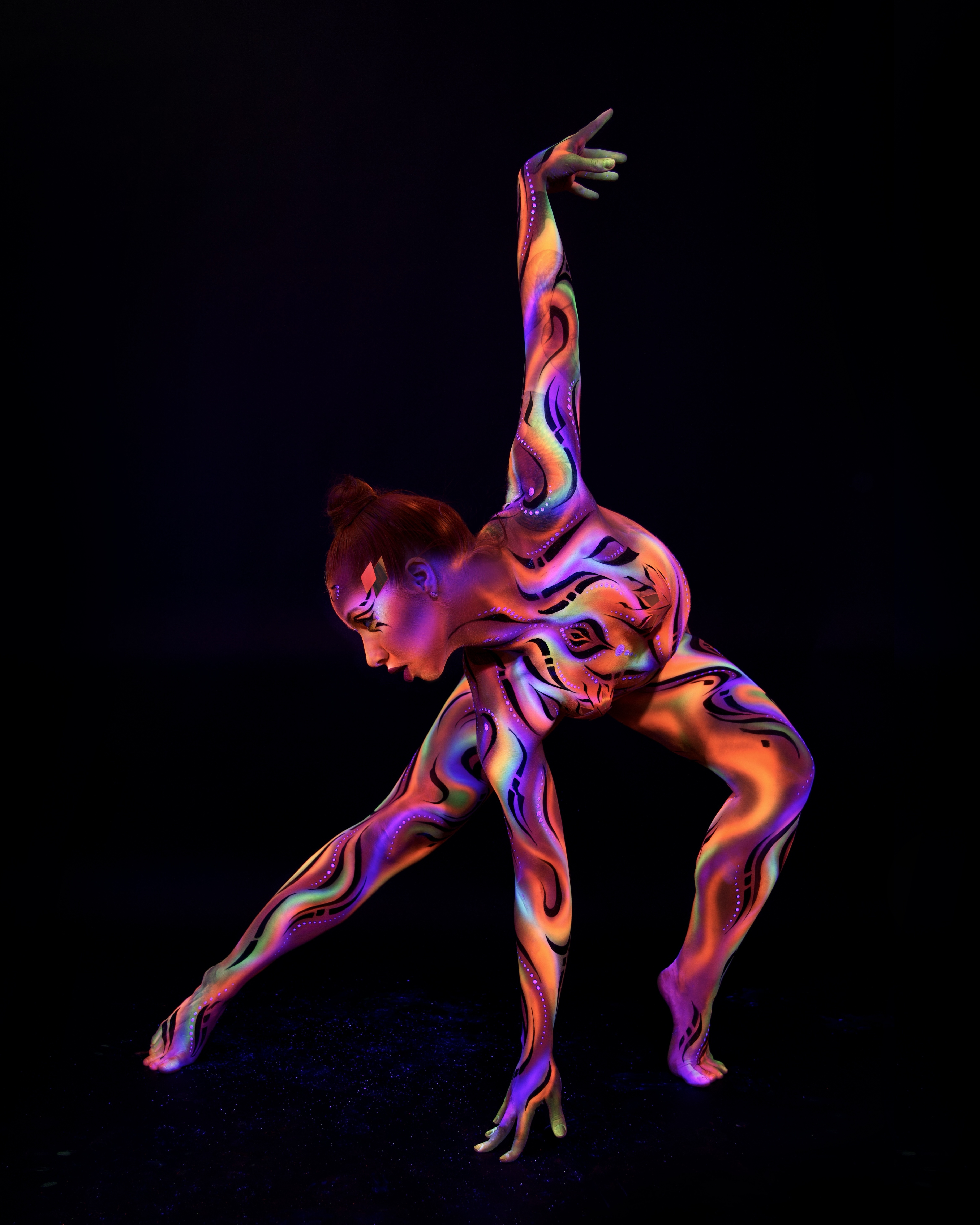Irina is photographed bent forward at the waist in a dramatic side-angled pose. The swirling marbled bodypaint in orange, purple and teal covers the torso and limbs, with a geometric pattern on the face. Bodypaint by Joana Rohr and Zami. Photographed by Elad Itzkin Photography under UV light in his studio in Gesundbrunnen, Berlin.