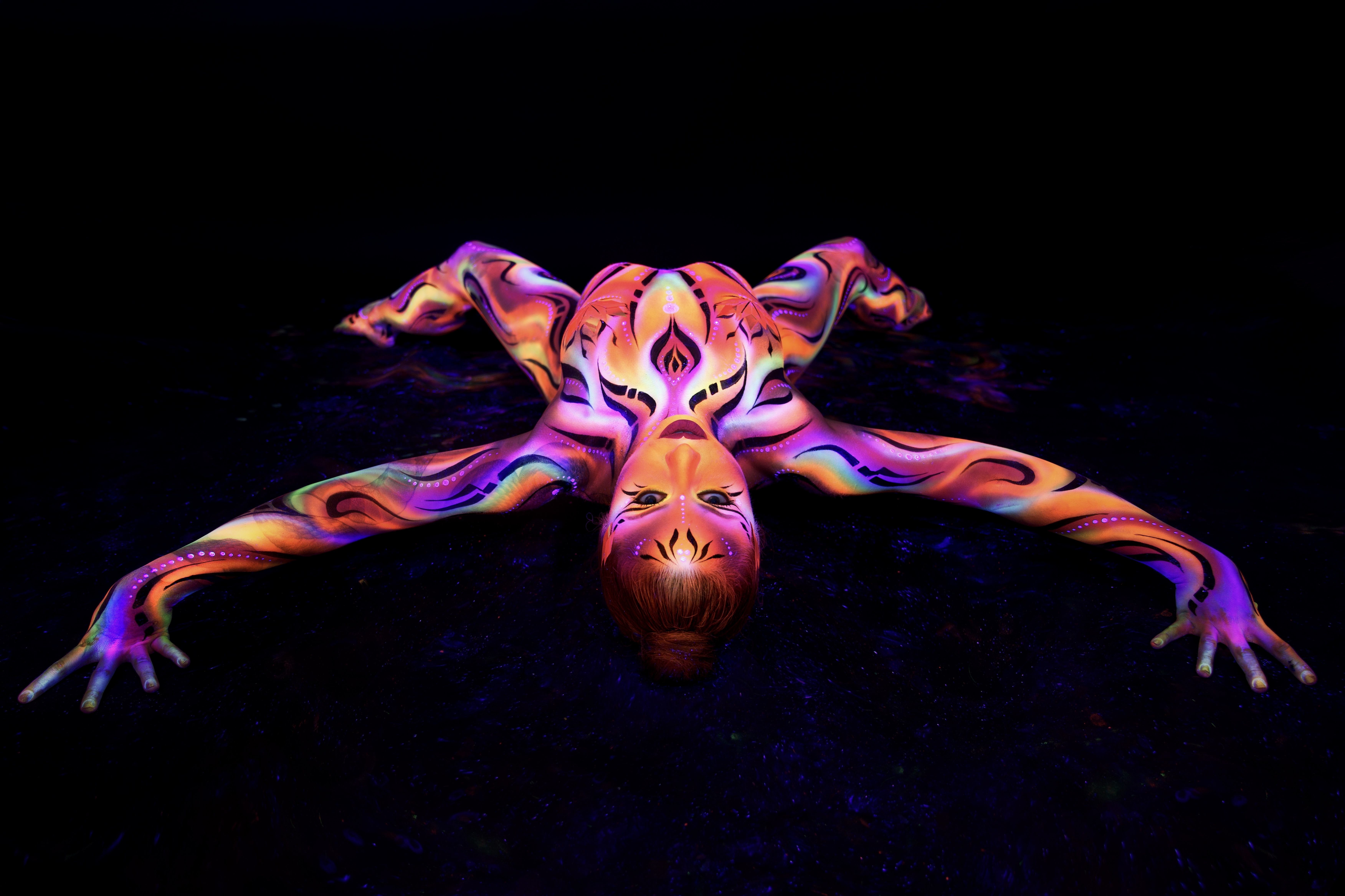 Irina lies on the floor with the body spread outward and the face toward the camera. The bodypaint is a marbled swirl pattern in orange, purple and pink covering the entire body, with a geometric design on the face. Bodypaint by Joana Rohr and Zami. Photographed by Elad Itzkin Photography under UV light in his studio in Gesundbrunnen, Berlin.