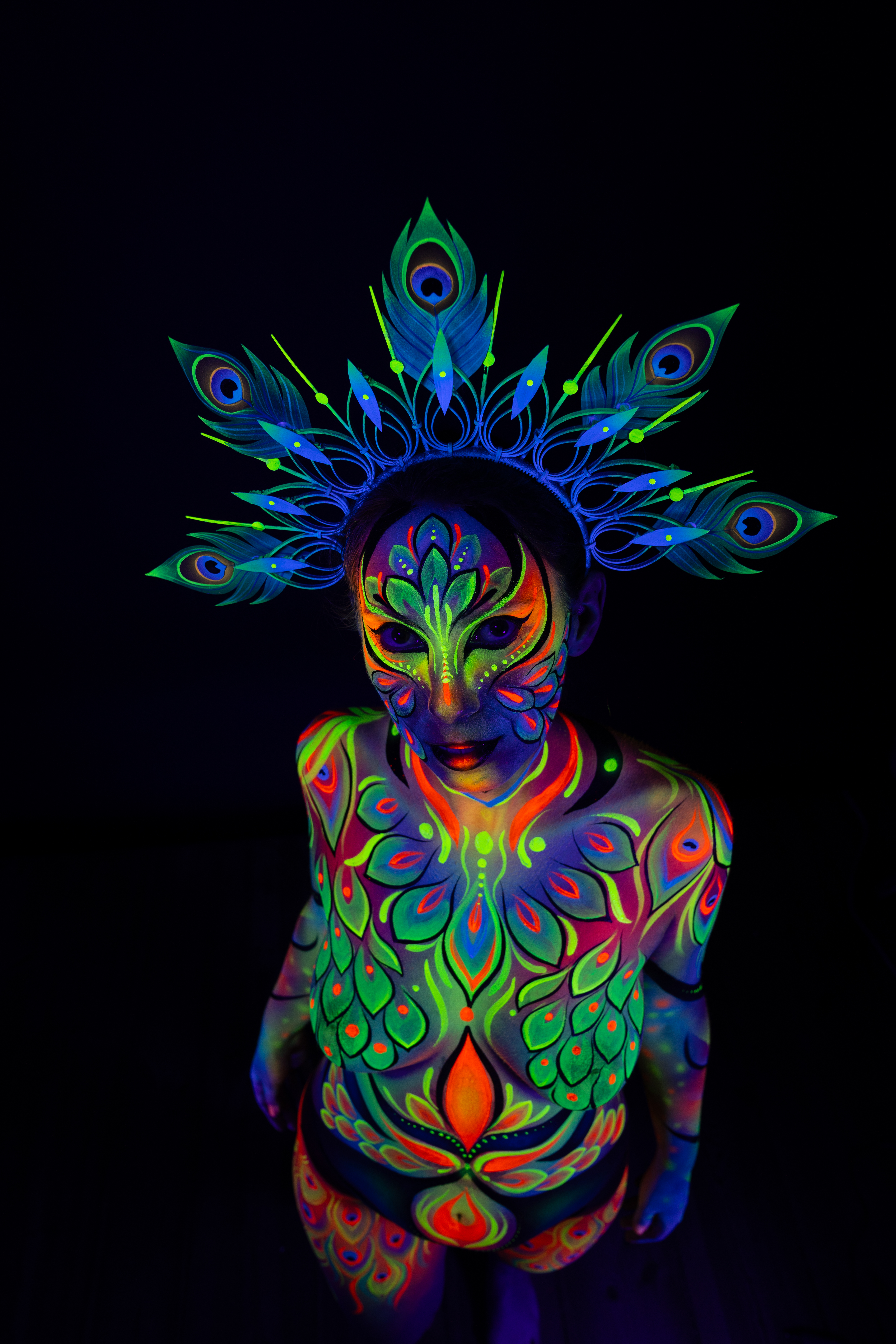 Alessia stands facing the camera with the head tilted downward, wearing a peacock feather themed crown. The bodypaint uses orange, green and blue tones in a symmetrical feathered pattern across the torso. Bodypaint by Joana Rohr. Photographed by Elad Itzkin Photography under UV light in his Berlin studio.