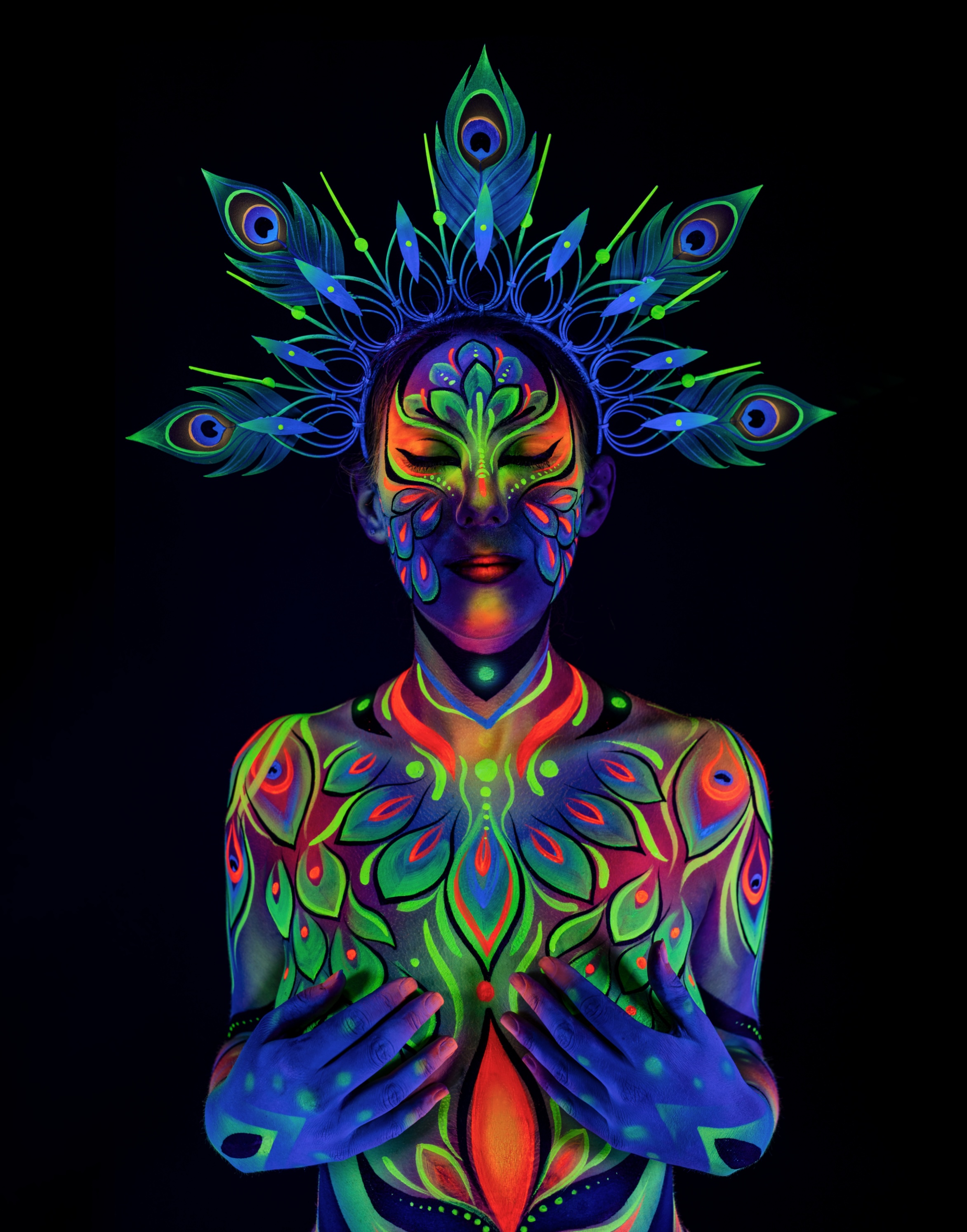 Alessia stands facing the camera beneath a peacock feather crown. The bodypaint design in orange, green and blue covers the chest and shoulders in a symmetrical feathered pattern. Bodypaint by Joana Rohr. Photographed by Elad Itzkin Photography under UV light in his studio in Gesundbrunnen, Berlin.