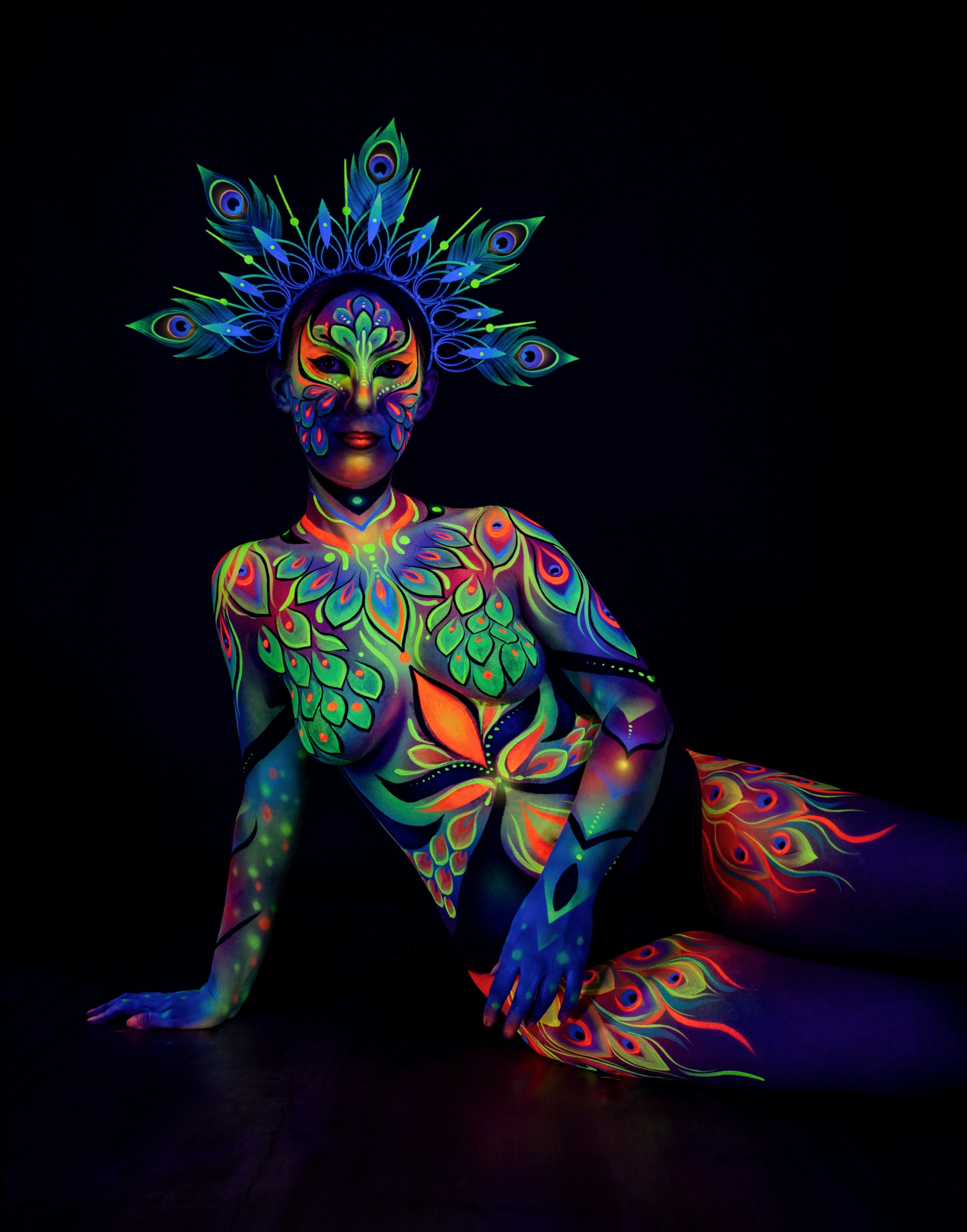 Alessia is seated on the floor with one leg extended, wearing a peacock feather crown. The orange, green and blue bodypaint design continues from the torso down along the legs. Bodypaint by Joana Rohr. Photographed by Elad Itzkin Photography under UV light in his Berlin studio.
