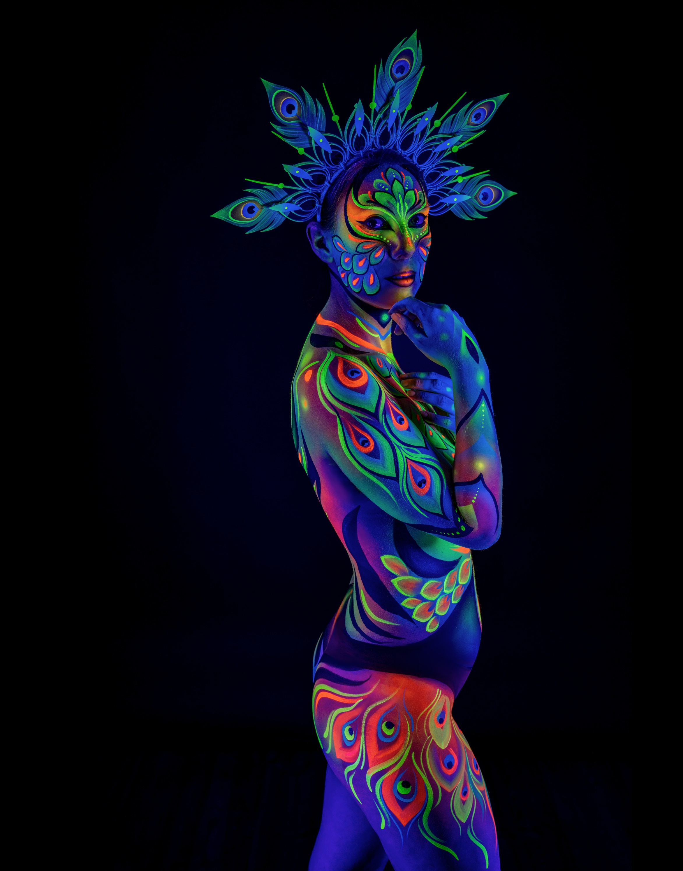 Alessia stands in a three-quarter side pose beneath a peacock feather crown. The bodypaint in orange, green and blue covers the torso, hip and leg in a feathered pattern. Bodypaint by Joana Rohr. Photographed by Elad Itzkin Photography under UV light in his studio in Gesundbrunnen, Berlin.