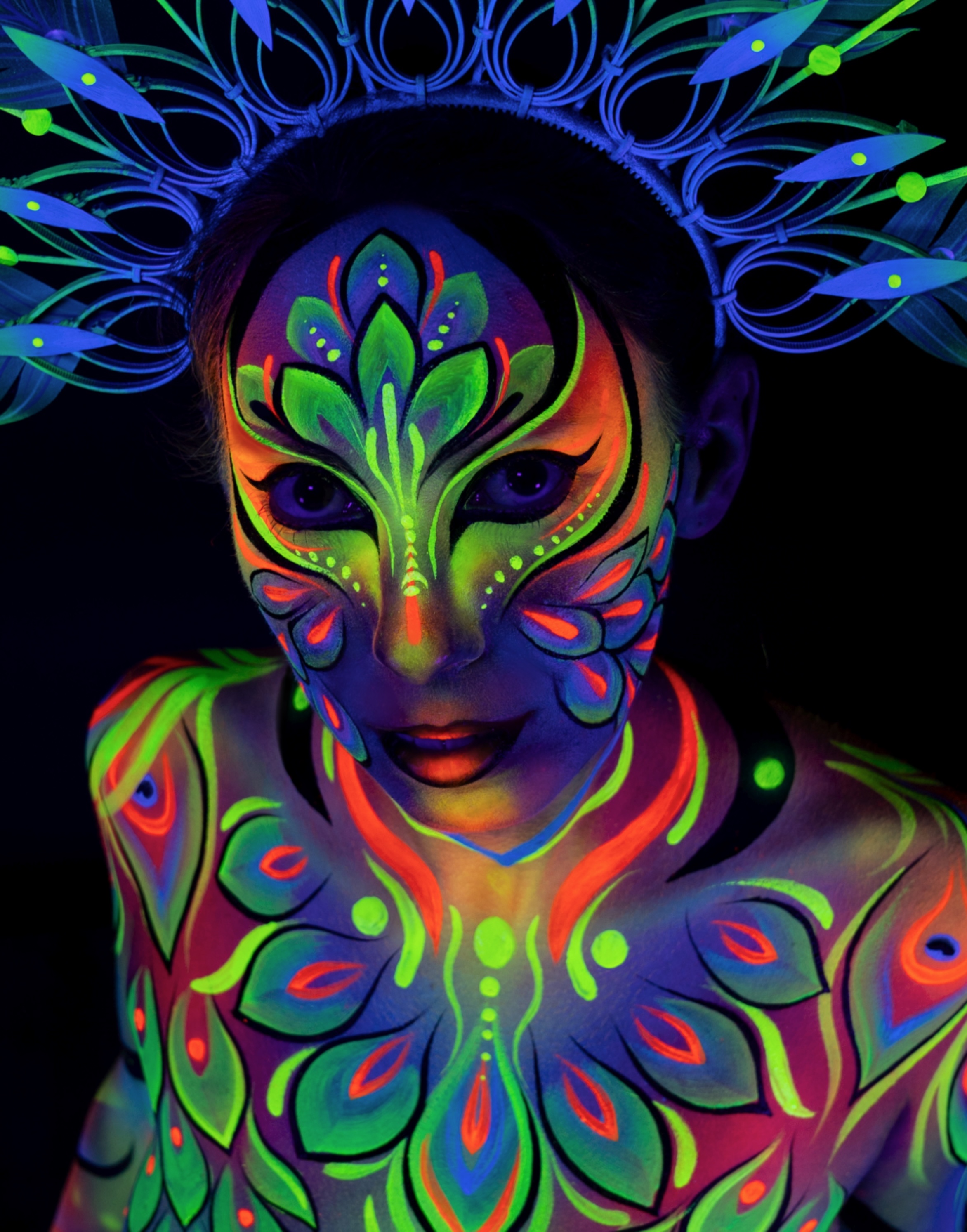 Alessia is shown from the chest up, facing the camera beneath a peacock feather crown. The bodypaint on the neck and shoulders uses orange, green and blue tones in a symmetrical pattern. Bodypaint by Joana Rohr. Photographed by Elad Itzkin Photography under UV light in his Berlin studio.