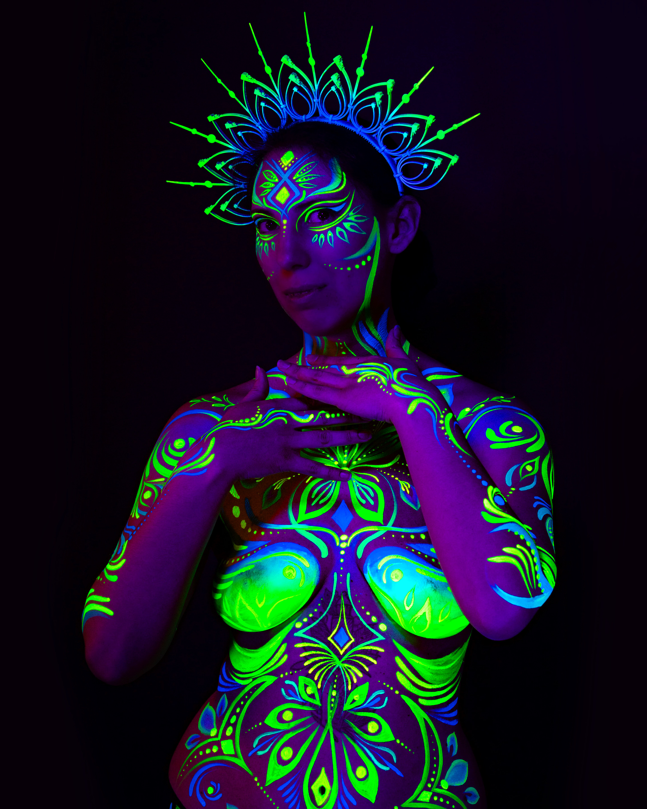 Alessia stands facing the camera with the head tilted slightly, beneath a radiating crown headpiece. The bodypaint design spans the torso in green and blue floral and leaf motifs. Bodypaint by Joana Rohr. Photographed by Elad Itzkin Photography under UV light in his studio in Gesundbrunnen, Berlin.