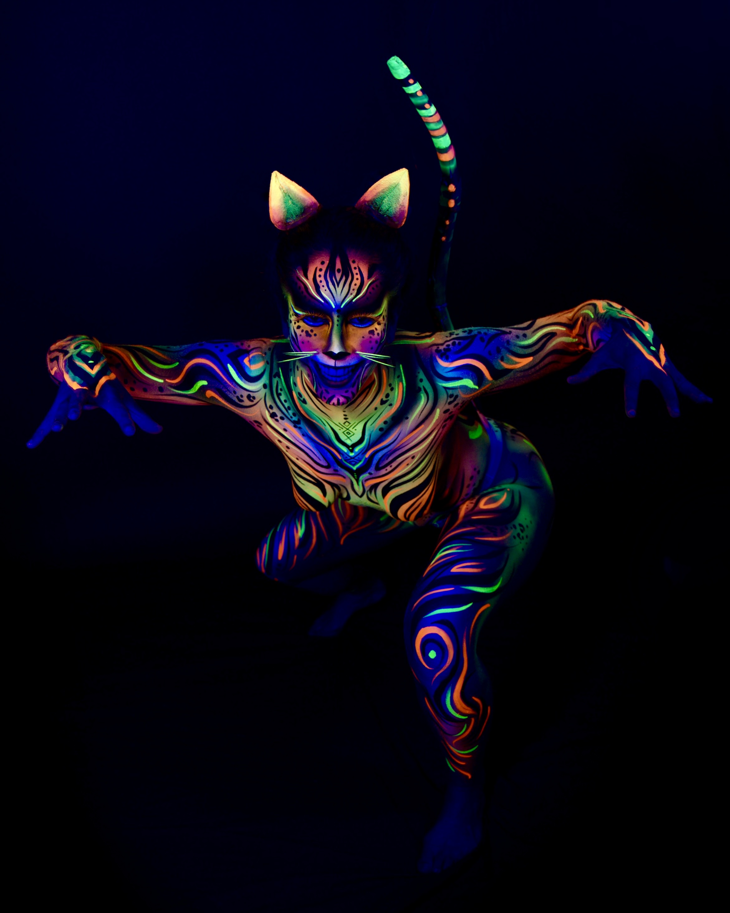 Chelsea is photographed crouching low in a dynamic pose, with a long painted tail curling upward behind the head. The cat themed bodypaint uses orange, blue and green tones in a wavy, striped pattern, with cat ear prosthetics. Bodypaint by Joana Rohr. Photographed by Elad Itzkin Photography under UV light in his studio in Gesundbrunnen, Berlin.