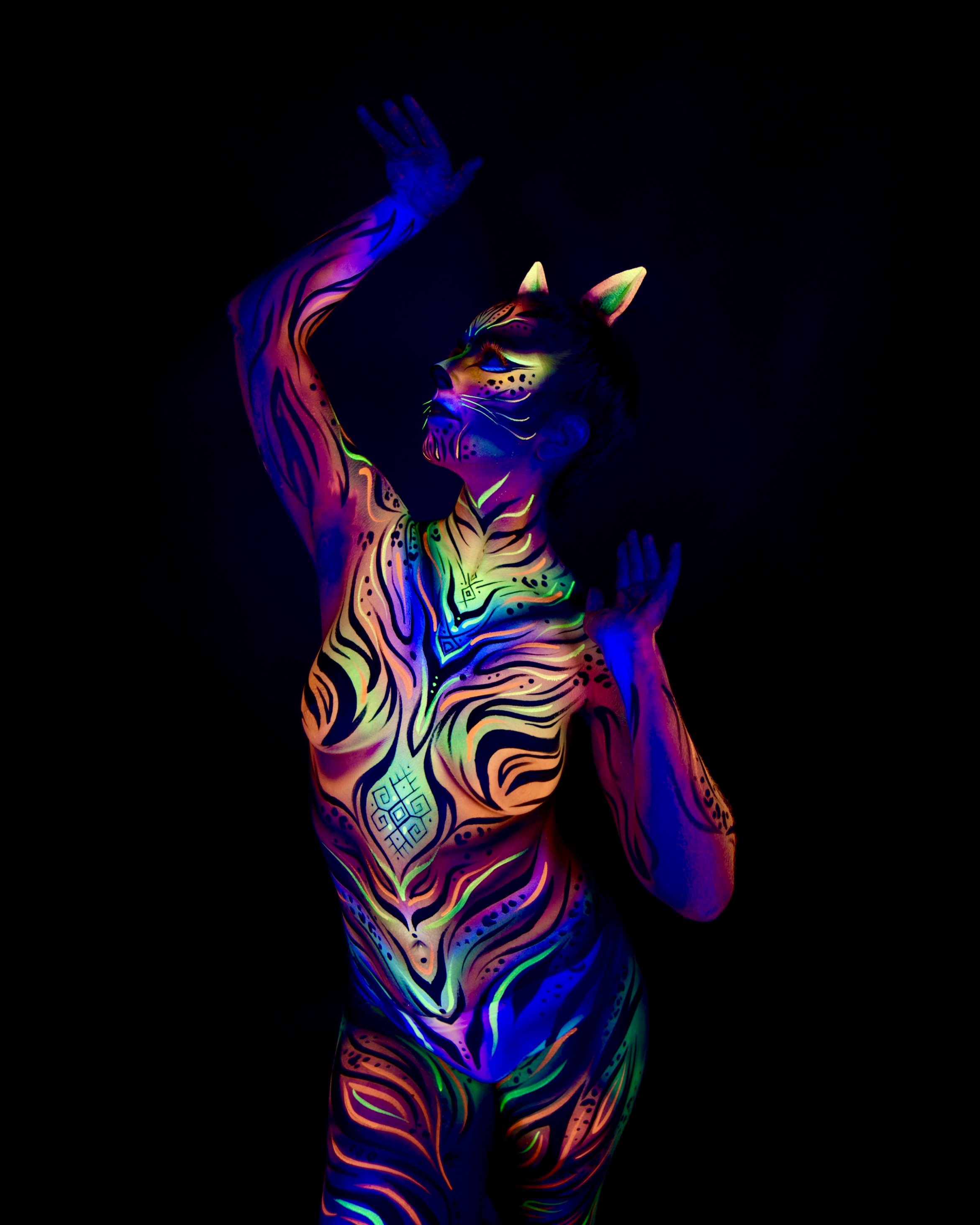 Chelsea stands with the head tilted back, wearing cat ear prosthetics as part of the design. The bodypaint covers the torso and legs in orange, blue and green wavy, striped linework. Bodypaint by Joana Rohr. Photographed by Elad Itzkin Photography under UV light in his Berlin studio.