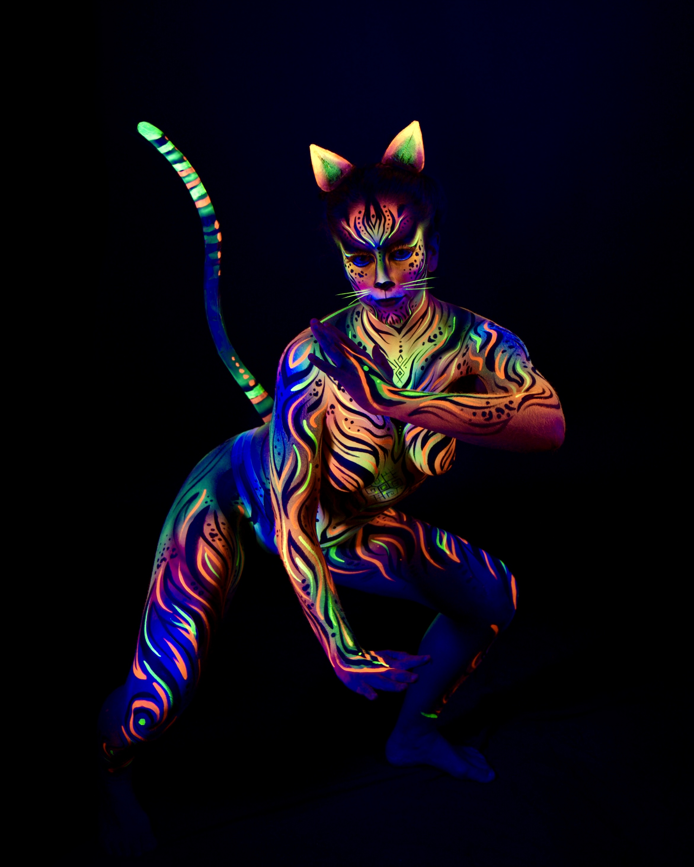 Chelsea crouches low with a long painted tail rising behind the head and cat ear prosthetics completing the look. The cat themed bodypaint uses orange, blue and green tones in a wavy striped pattern. Bodypaint by Joana Rohr. Photographed by Elad Itzkin Photography under UV light in his studio in Gesundbrunnen, Berlin.