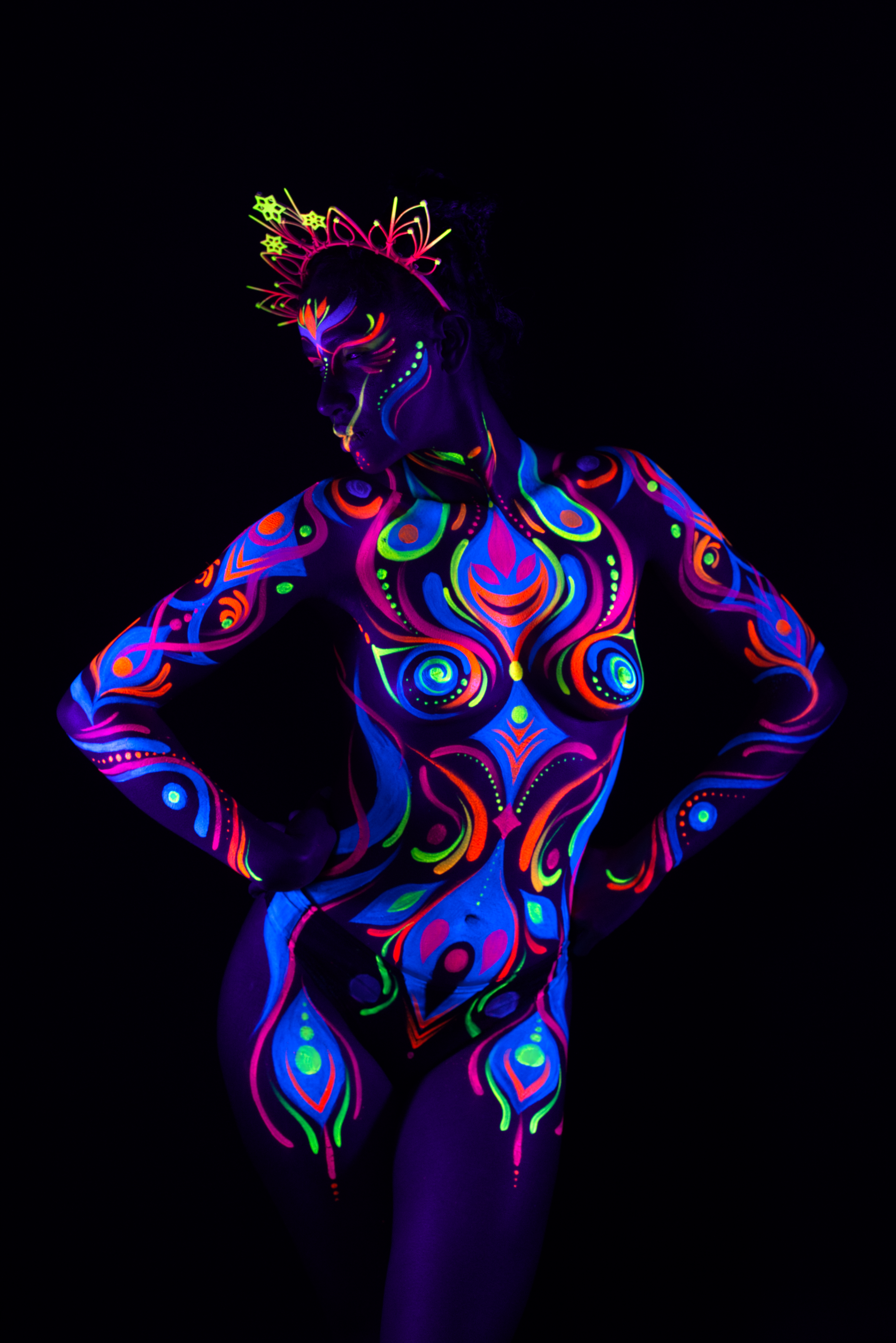 Lydia stands facing the camera beneath a radiating crown headpiece. The bodypaint is a swirl and paisley pattern in pink, blue, orange and green spanning the torso. Bodypaint by Joana Rohr. Photographed by Elad Itzkin Photography under UV light in his studio in Gesundbrunnen, Berlin.