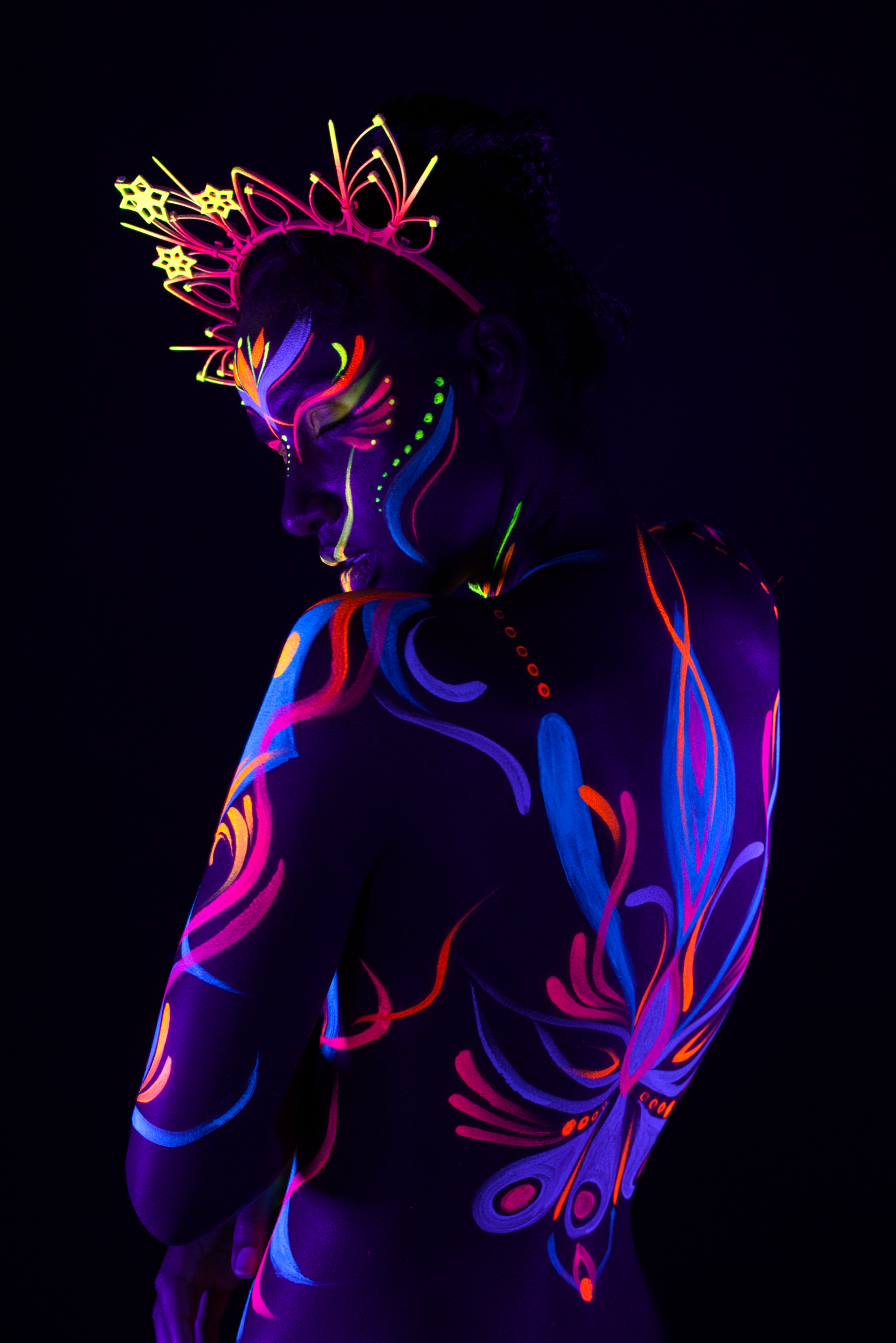 Lydia stands in a three-quarter pose with the head turned downward, wearing a radiating crown headpiece. The swirling bodypaint design in pink, blue and orange covers the torso and shoulder. Bodypaint by Joana Rohr. Photographed by Elad Itzkin Photography under UV light in his studio in Gesundbrunnen, Berlin.