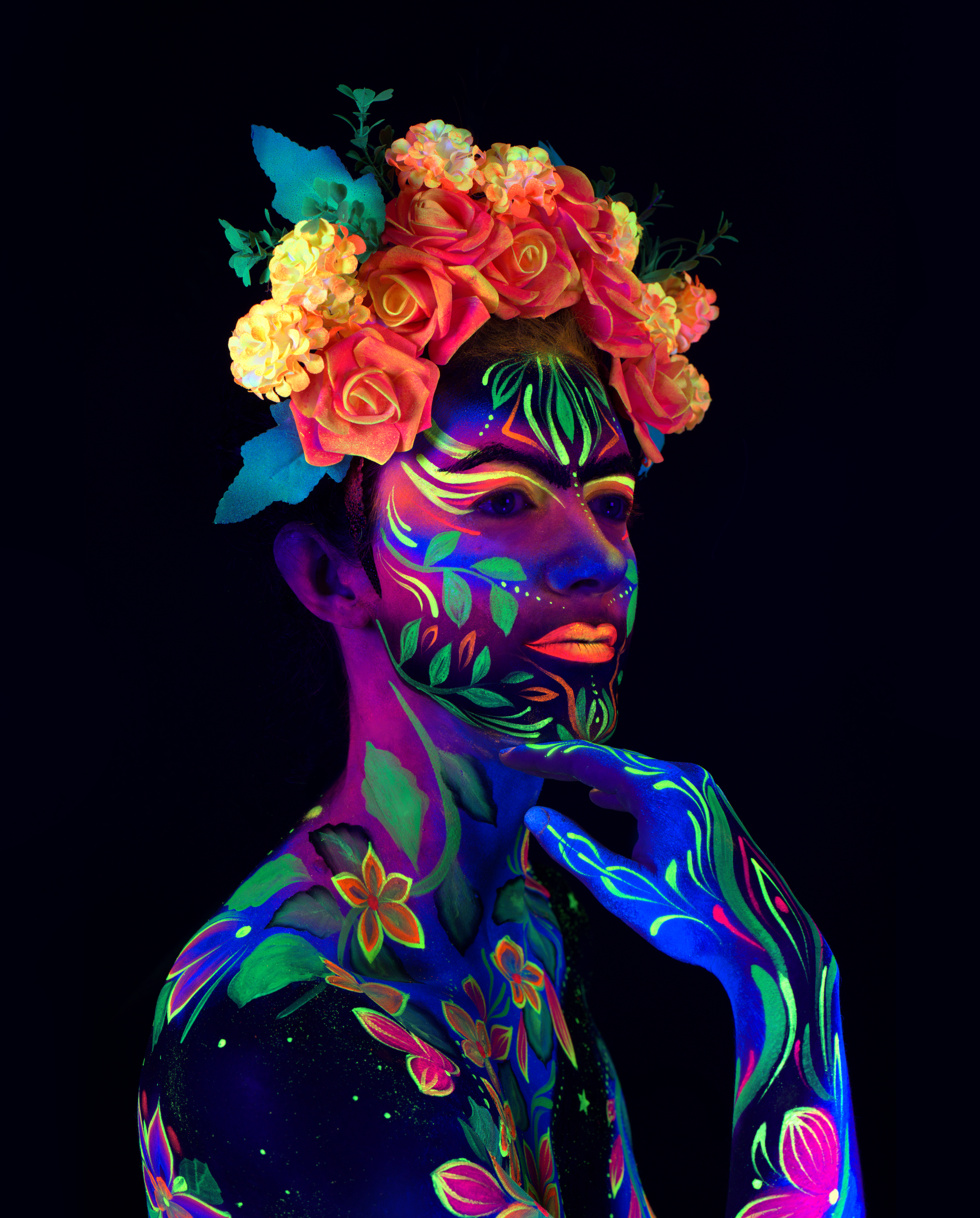 Maria is shown from the shoulders up wearing a floral crown headpiece. The bodypaint on the face and shoulders is a floral design in green and orange with leaf and blossom motifs. Bodypaint by Joana Rohr. Photographed by Elad Itzkin Photography under UV light in his Berlin studio.