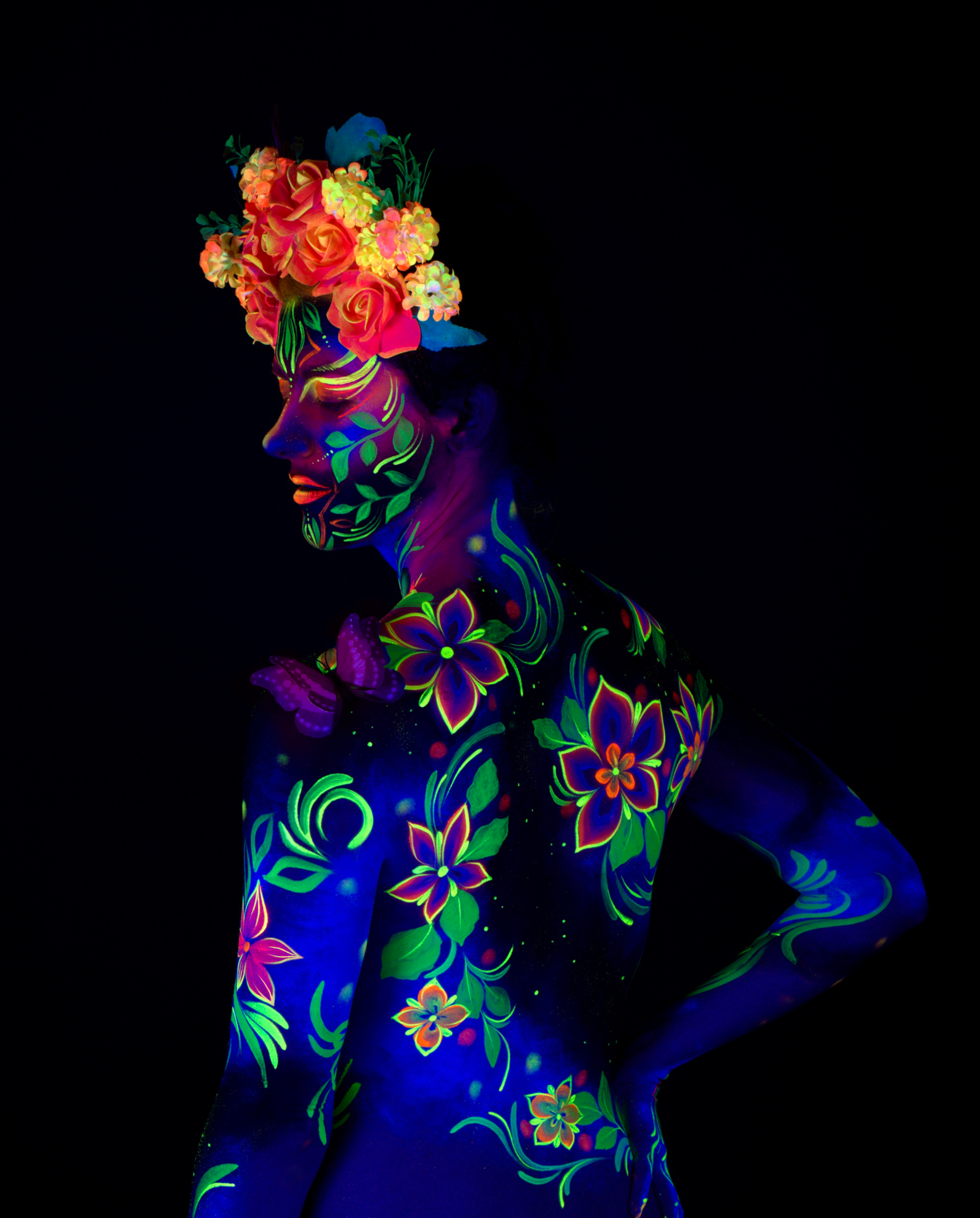 Maria stands in a three-quarter side pose wearing a floral crown headpiece. The bodypaint covers the torso and shoulder in a floral design of blue and green with pink and orange blossoms. Bodypaint by Joana Rohr. Photographed by Elad Itzkin Photography under UV light in his studio in Gesundbrunnen, Berlin.