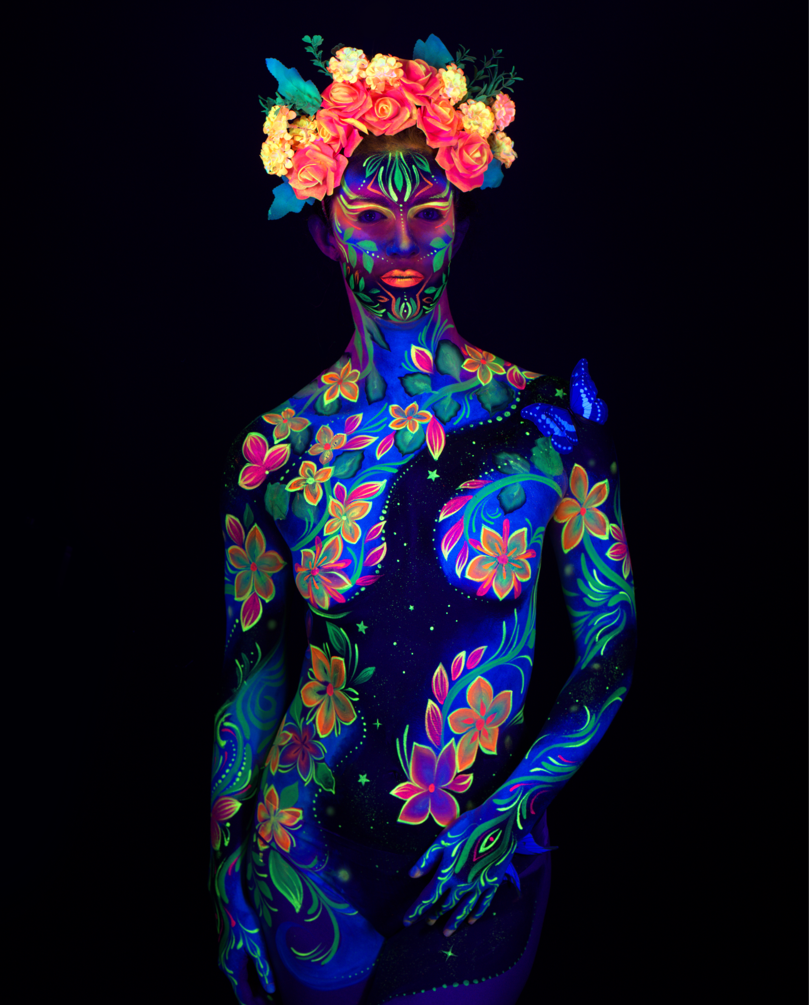 Maria stands in a three-quarter pose facing the camera, wearing a floral crown headpiece. Her body is painted in an orange, pink, and green floral pattern over a deep blue base, covering the torso and limbs. Bodypaint by Joana Rohr. Photographed by Elad Itzkin Photography under UV light in his studio in Gesundbrunnen, Berlin.