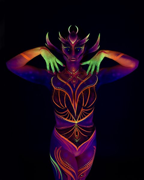 Rala du Reign stands facing the camera in an upright pose. The design is a purple and orange tribal-style pattern with a horned, mask-like face design and abstract linework across the chest and torso. Bodypaint by Joana Rohr. Photographed by Elad Itzkin Photography under UV light in his studio in Gesundbrunnen, Berlin.