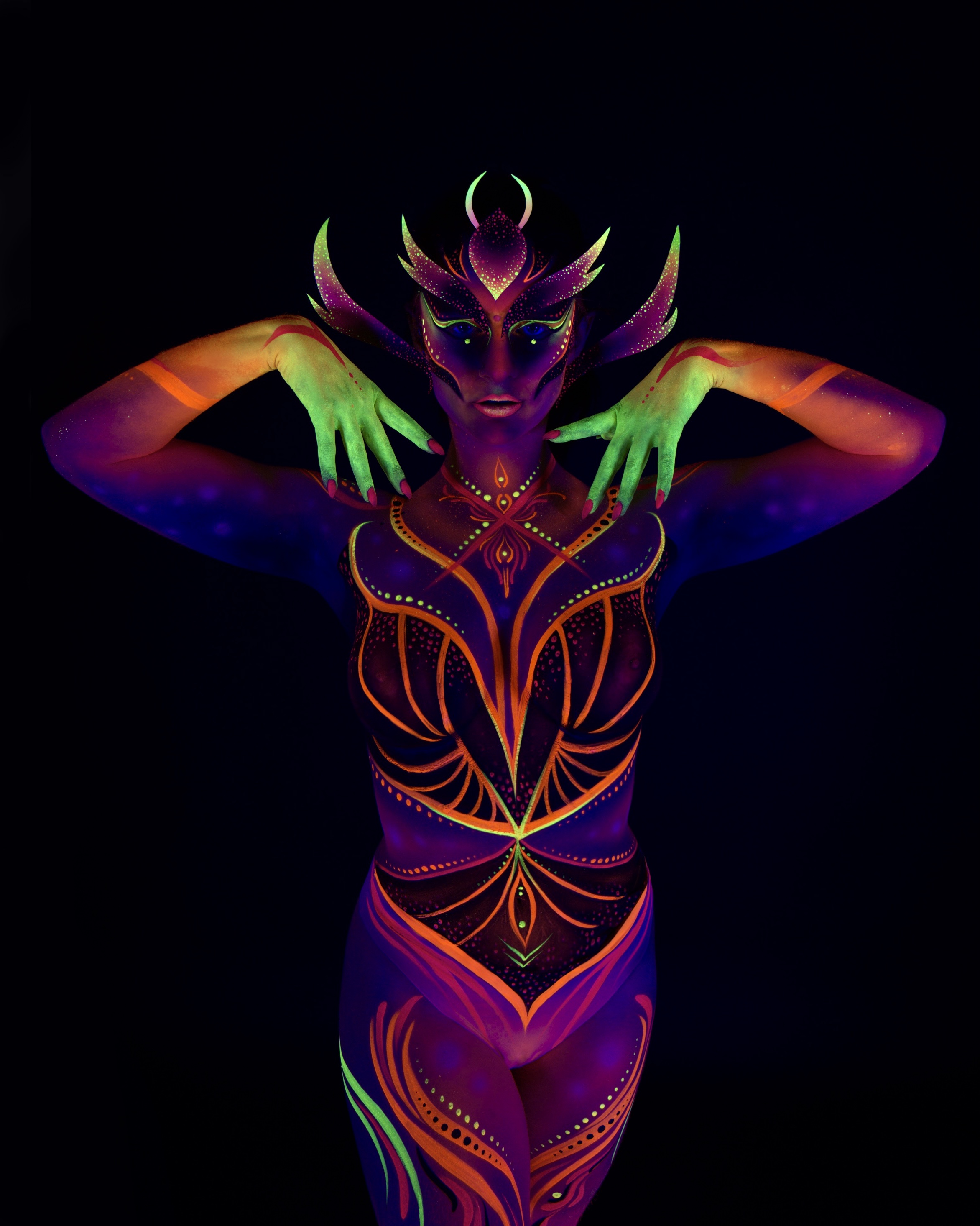 Rala du Reign stands facing the camera in an upright pose. The design is a purple and orange tribal-style pattern with a horned, mask-like face design and abstract linework across the chest and torso. Bodypaint by Joana Rohr. Photographed by Elad Itzkin Photography under UV light in his studio in Gesundbrunnen, Berlin.