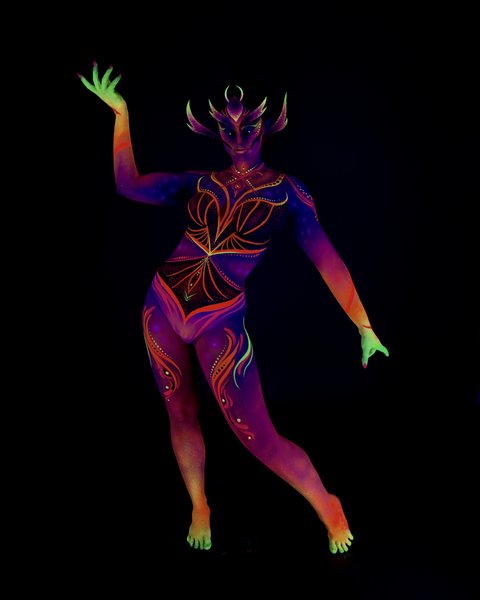 Rala du Reign stands in a dynamic three-quarter pose, leaning slightly forward. The bodypaint is a purple and orange tribal-style design with a horned, mask-like face pattern and abstract linework across the torso and legs. Bodypaint by Joana Rohr. Photographed by Elad Itzkin Photography under UV light in his studio in Gesundbrunnen, Berlin.