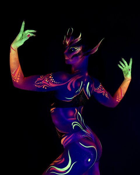 Rala du Reign is photographed with her back to the camera, upper body twisted to show a side profile of the face. The purple and orange tribal-style design continues across the back and torso, with a horned, mask-like face pattern. Bodypaint by Joana Rohr. Photographed by Elad Itzkin Photography under UV light in his studio in Gesundbrunnen, Berlin.