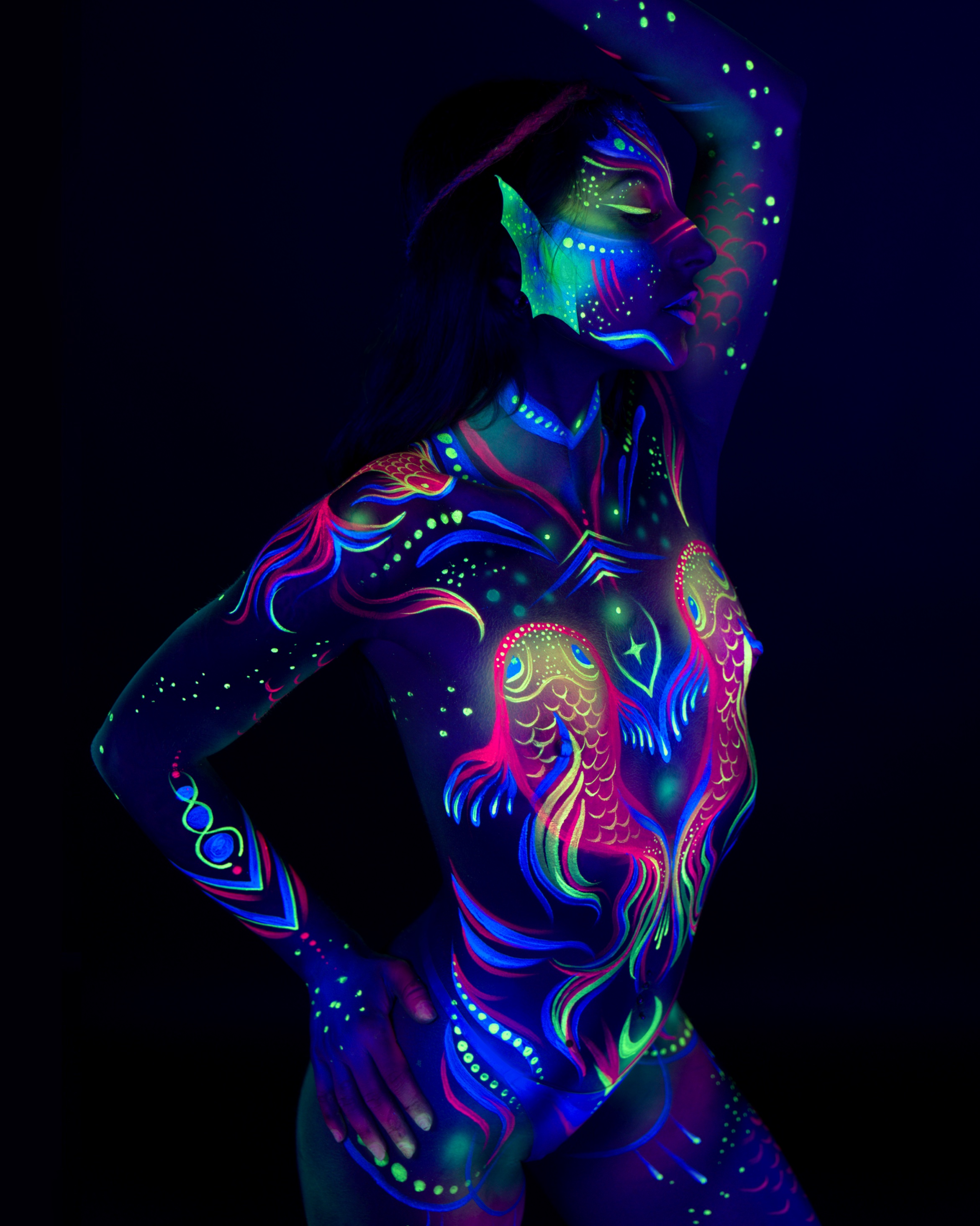 Sori stands in a side three-quarter pose, body turned toward the camera. Her torso is painted with a pink, blue, and green fish motif, along with a mask-like face design and fin-shaped detail at the ear. Bodypaint by Joana Rohr. Photographed by Elad Itzkin Photography under UV light in his studio in Gesundbrunnen, Berlin.