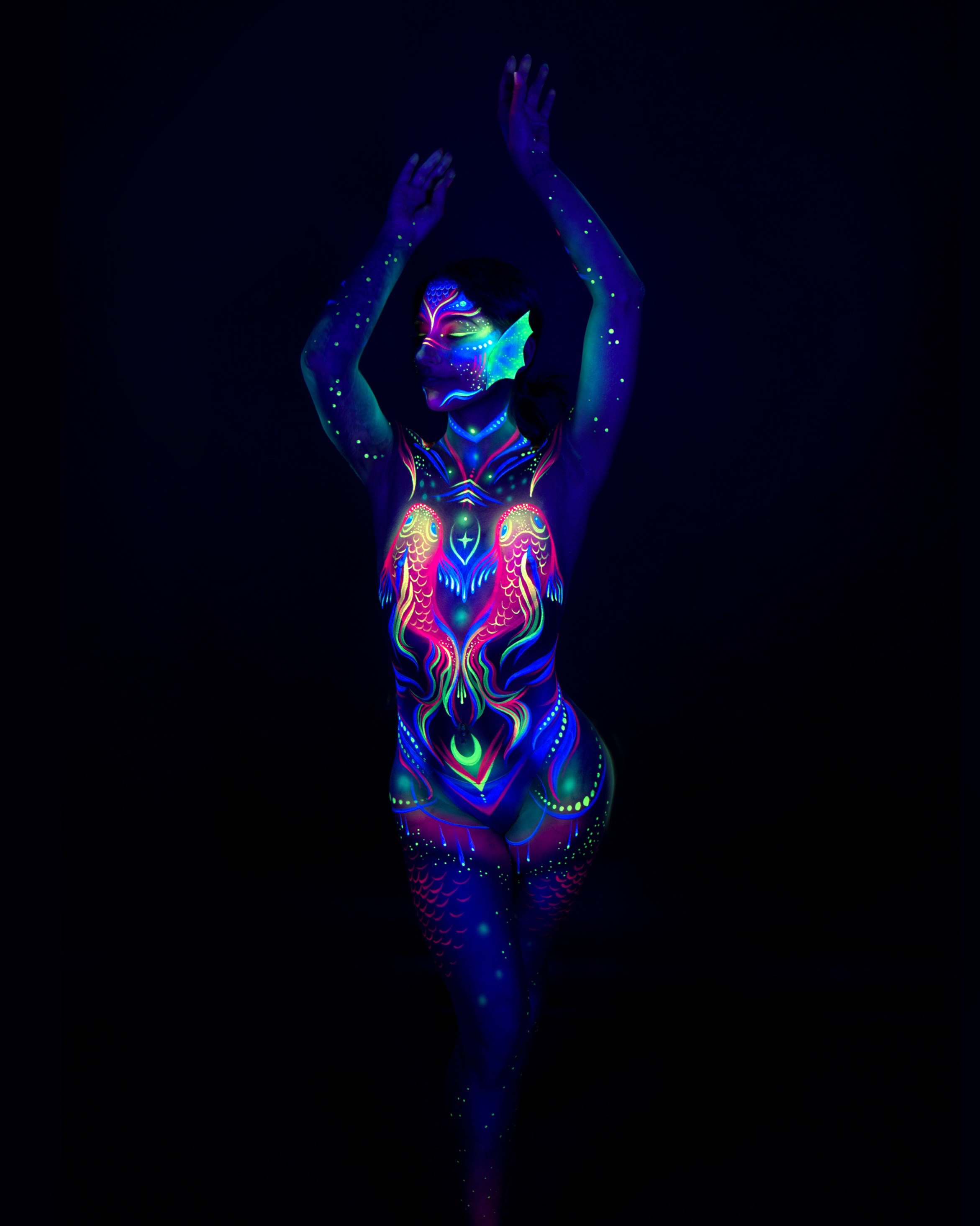 Sori stands facing the camera in an upright pose. The bodypaint design shows a pink, blue, and green abstract pattern across the chest, with a mask-like face design. Bodypaint by Joana Rohr. Photographed by Elad Itzkin Photography under UV light in his studio in Gesundbrunnen, Berlin.