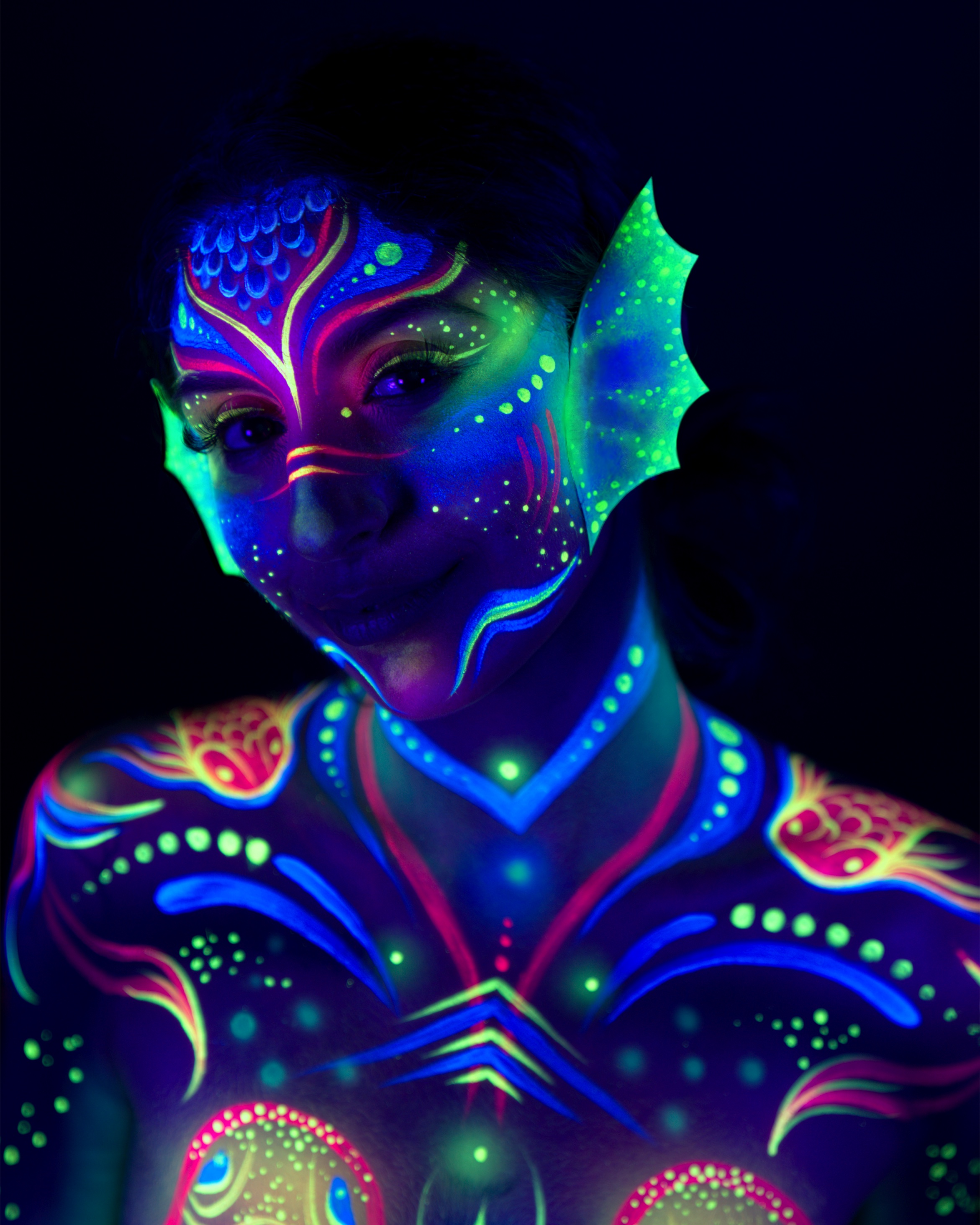 Sori is shown in a close-up, upper-body portrait facing the camera directly. The design includes a mask-like face pattern in blue and green with a fin-shaped detail near the ear, and a pink, blue, and green fish motif visible on the chest. Bodypaint by Joana Rohr. Photographed by Elad Itzkin Photography under UV light in his studio in Gesundbrunnen, Berlin.
