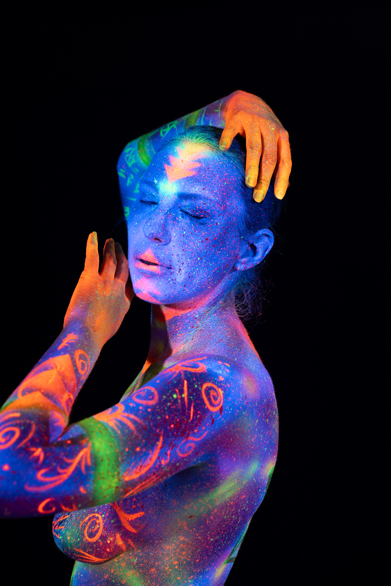 Alessia stands turned to one side in a three-quarter pose. Orange, green and pink swirling linework covers her shoulder and upper torso over a blue glitter base. Bodypaint by Elad Itzkin, Geli. Photographed by Elad Itzkin Photography under UV light in his Gesundbrunnen, Berlin studio.