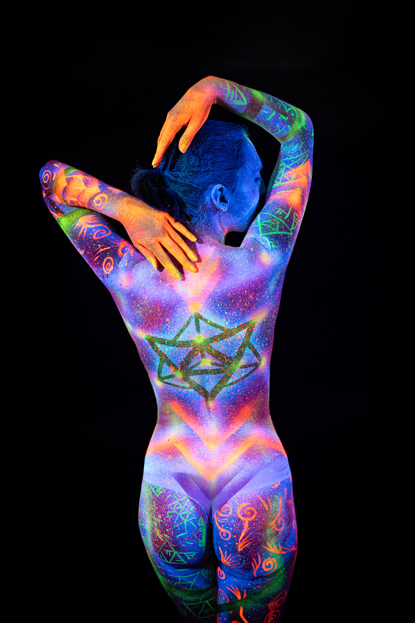 Alessia is photographed from behind, her head turned in profile. A green and orange geometric star design glows across her back over a purple and blue UV base. Bodypaint by Elad Itzkin, Geli. Photographed by Elad Itzkin Photography under blacklight in his Gesundbrunnen, Berlin studio.