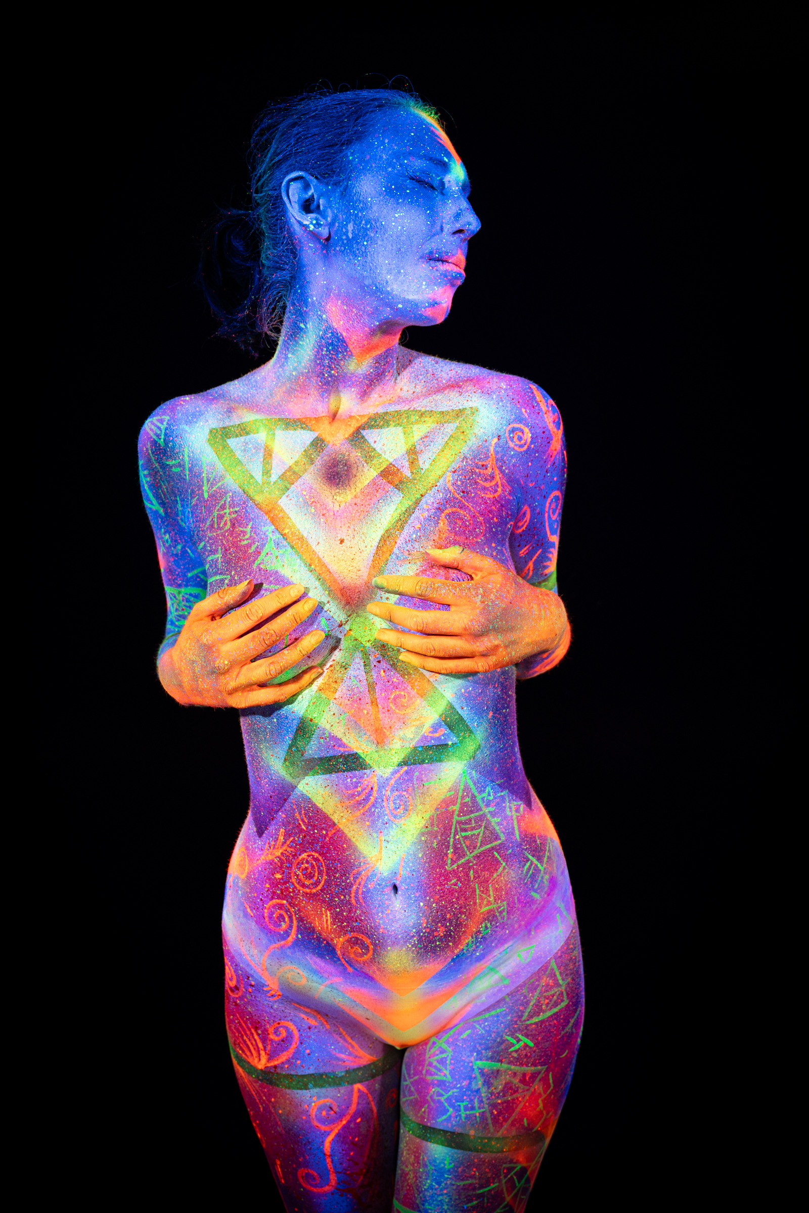Alessia stands with her head turned slightly to the side. Her torso features a layered triangular geometric design in green and orange over a blue and purple UV base. Bodypaint by Elad Itzkin, Geli. Photographed by Elad Itzkin Photography under blacklight in his Gesundbrunnen, Berlin studio.