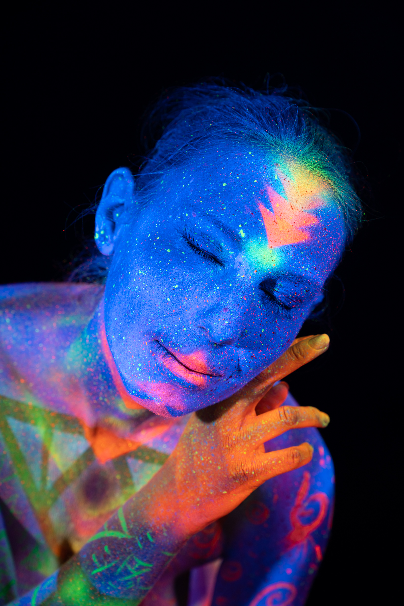 In this close portrait, Alessia's head is turned to one side. Her skin shows a blue UV base with an orange geometric accent on the forehead and scrollwork on the shoulder. Bodypaint by Elad Itzkin, Geli. Photographed by Elad Itzkin Photography under UV light in Gesundbrunnen, Berlin.