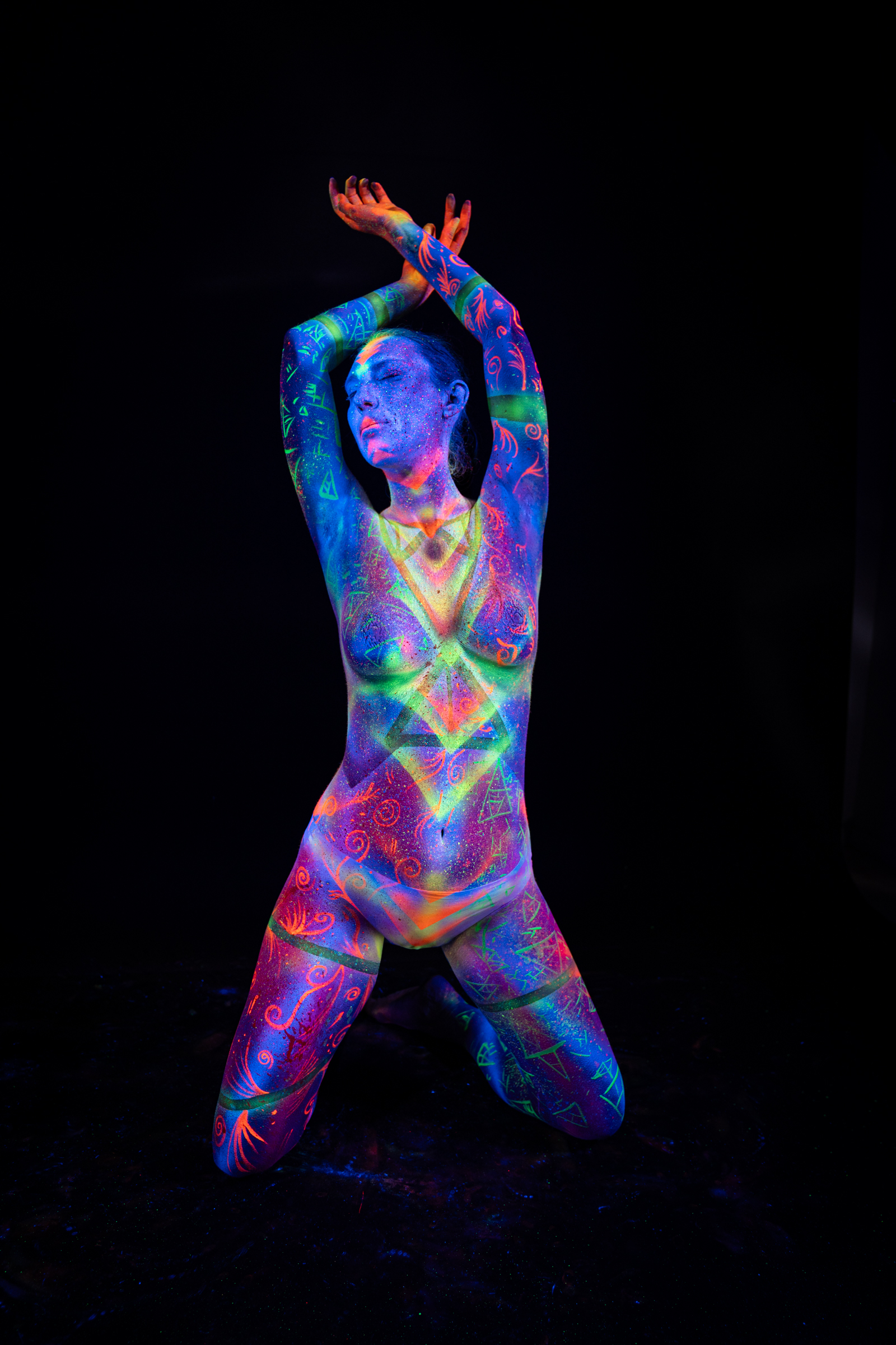 Alessia kneels upright on the floor. A symmetrical chevron and diamond geometric pattern in green, orange and pink covers her torso over a blue UV base. Bodypaint by Elad Itzkin, Geli. Photographed by Elad Itzkin Photography under UV light in his Gesundbrunnen, Berlin studio.