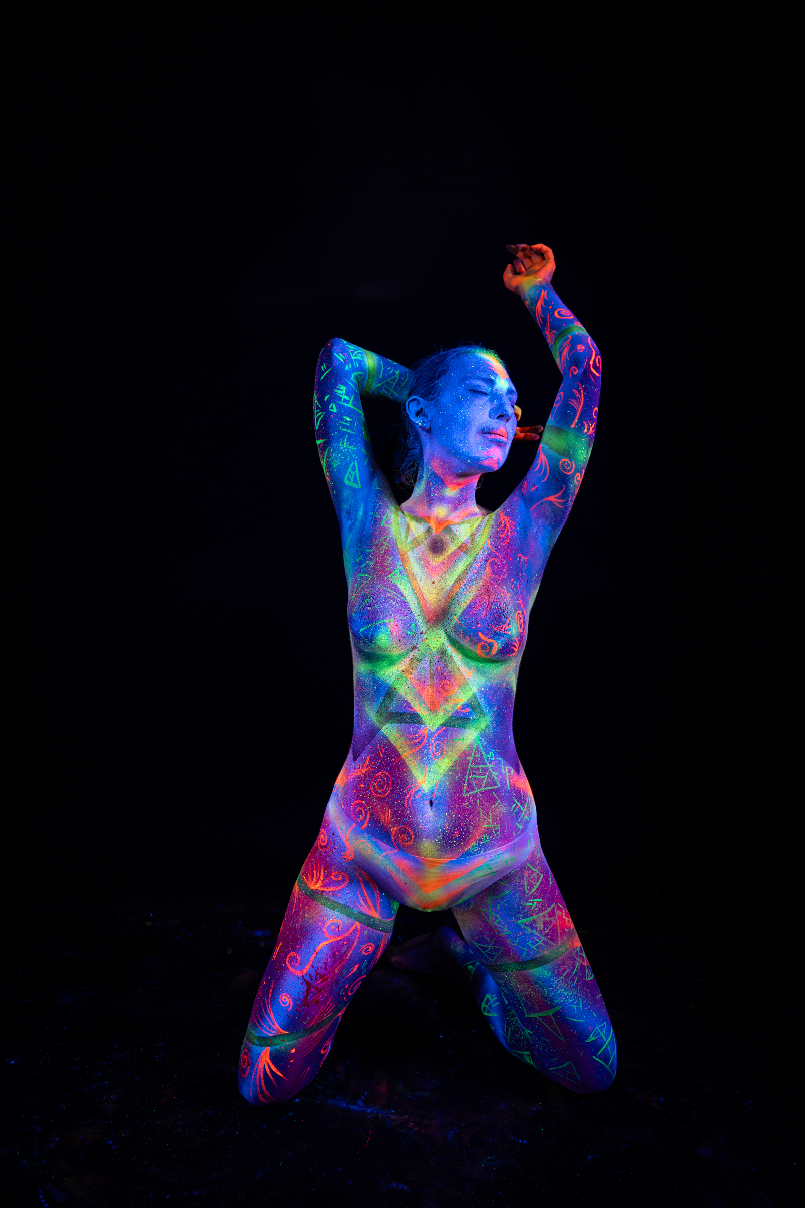 Alessia kneels facing the camera in an upright position. A triangular geometric design in green and orange covers her chest, set against scattered UV detail on a blue and purple base. Bodypaint by Elad Itzkin, Geli. Photographed by Elad Itzkin Photography under UV light in Gesundbrunnen, Berlin.