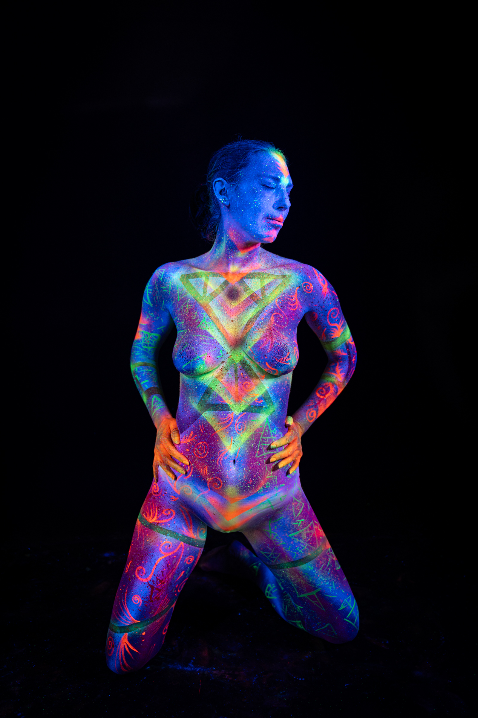 Alessia kneels facing forward in an upright pose. Her chest and abdomen display a layered triangular geometric motif in green and orange over a purple UV base. Bodypaint by Elad Itzkin, Geli. Photographed by Elad Itzkin Photography under blacklight in his Gesundbrunnen, Berlin studio.