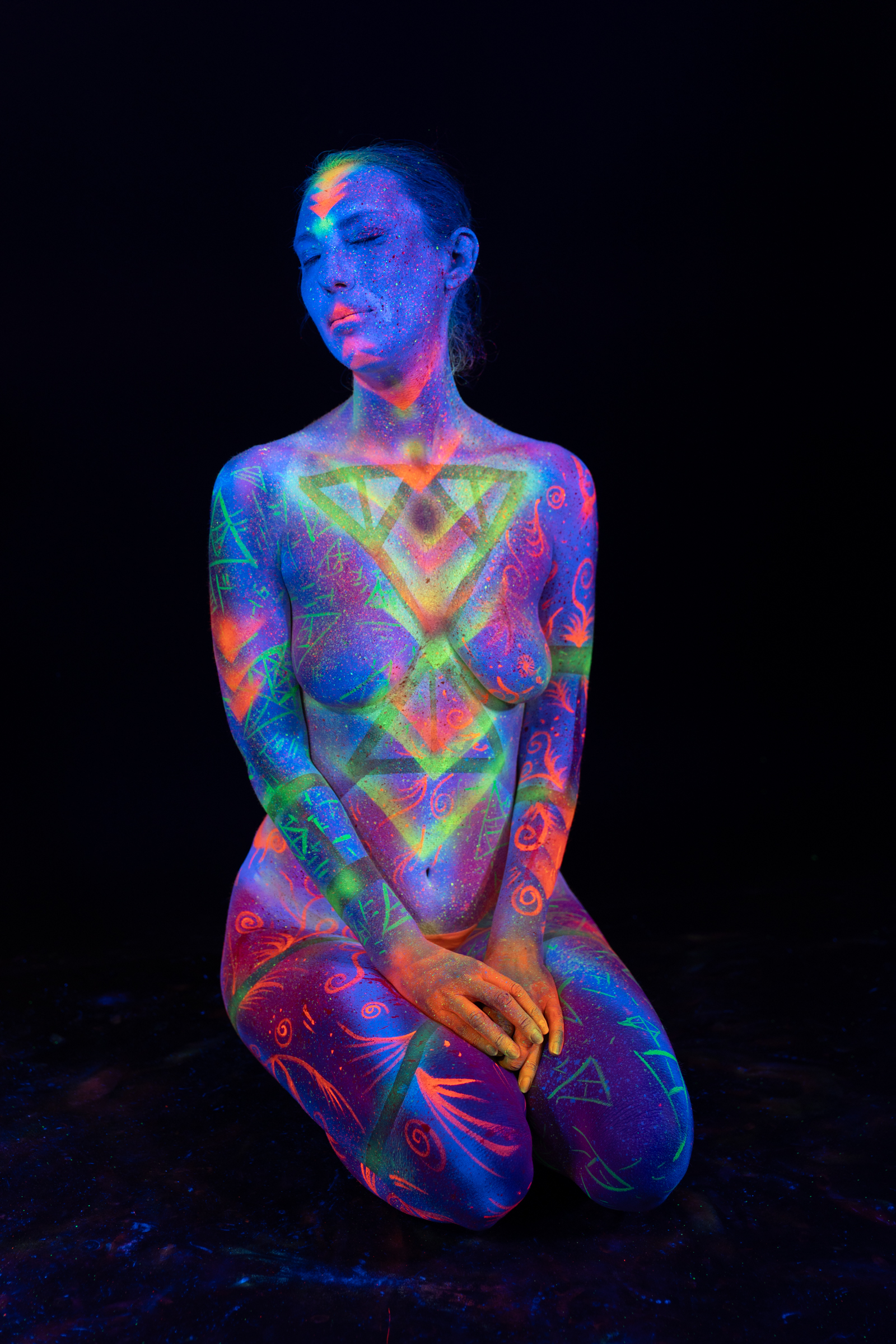 Alessia kneels facing the camera, her head tilted slightly forward. Her torso is painted with a symmetrical triangular design in green and orange over a blue and pink UV base. Bodypaint by Elad Itzkin, Geli. Photographed by Elad Itzkin Photography under UV light in Gesundbrunnen, Berlin.