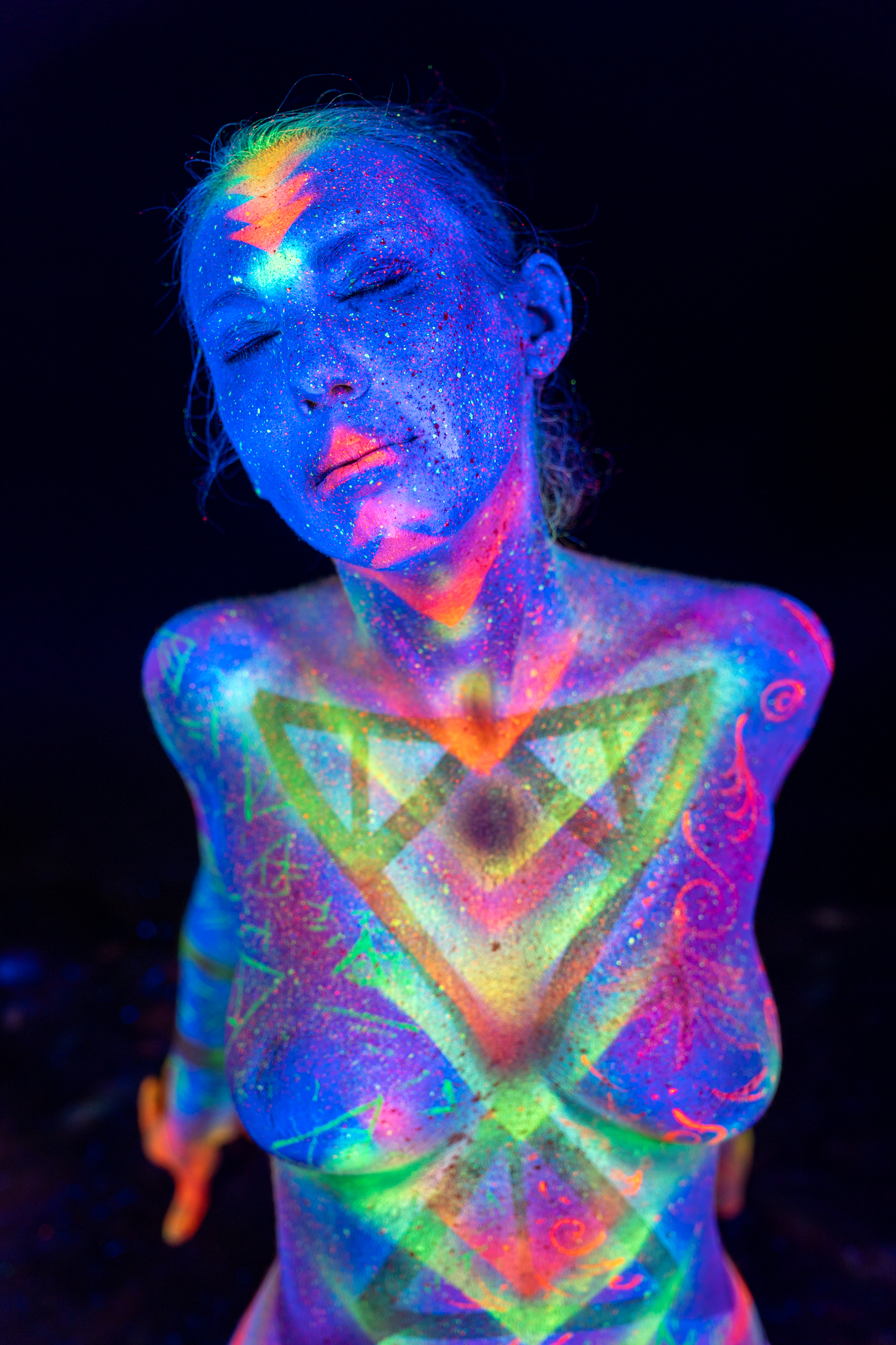Alessia stands facing the camera in an upper-body view, her head tilted slightly down. Her chest and shoulders show a triangular geometric UV design in green and orange over a blue base. Bodypaint by Elad Itzkin, Geli. Photographed by Elad Itzkin Photography under UV light in Gesundbrunnen, Berlin.