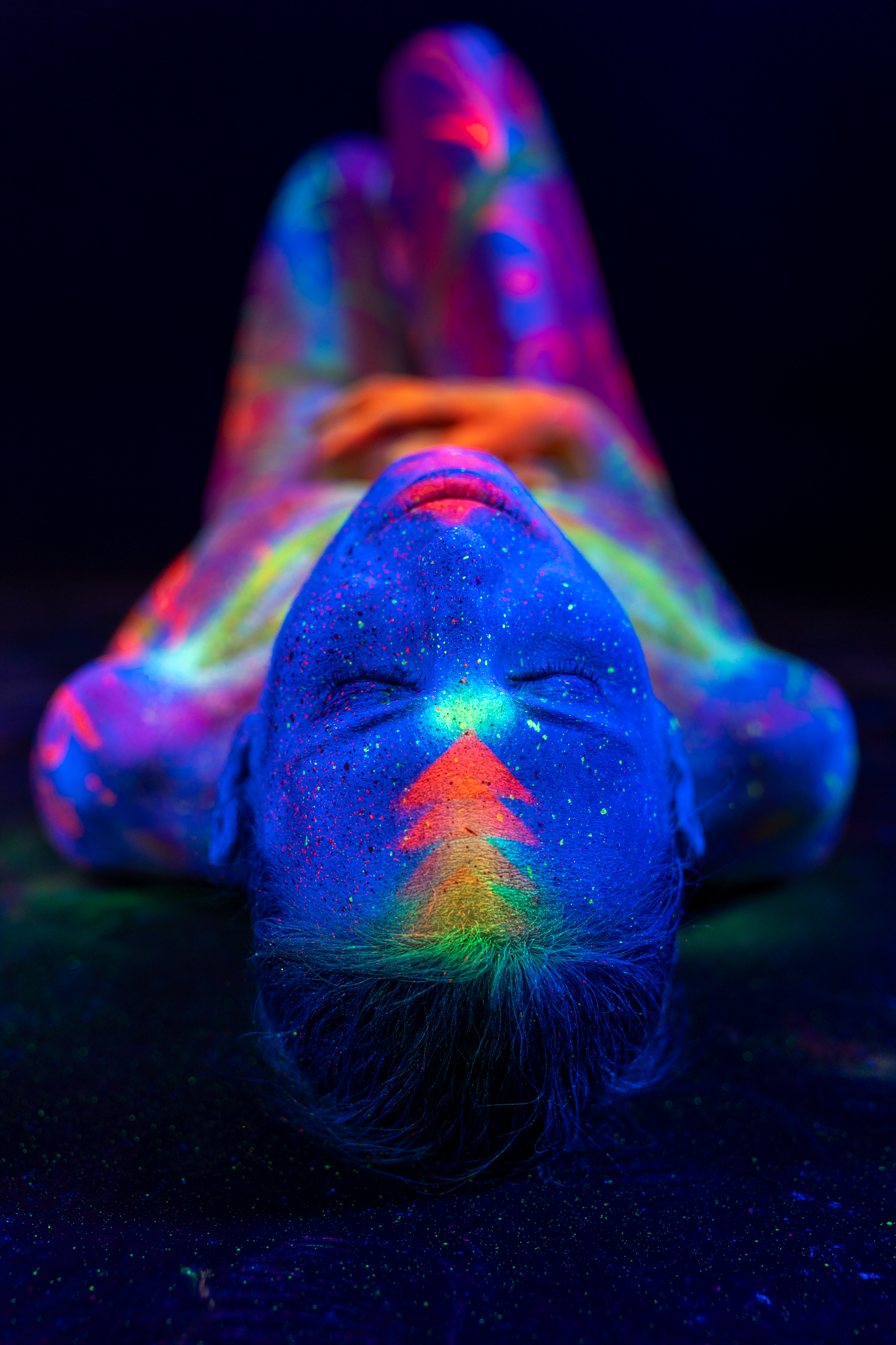Alessia is photographed lying down with her knees bent, her head toward the bottom of the frame. Her face carries a blue UV base with an orange geometric accent, and her legs show colorful scrollwork. Bodypaint by Elad Itzkin, Geli. Photographed by Elad Itzkin Photography under UV light in Gesundbrunnen, Berlin.