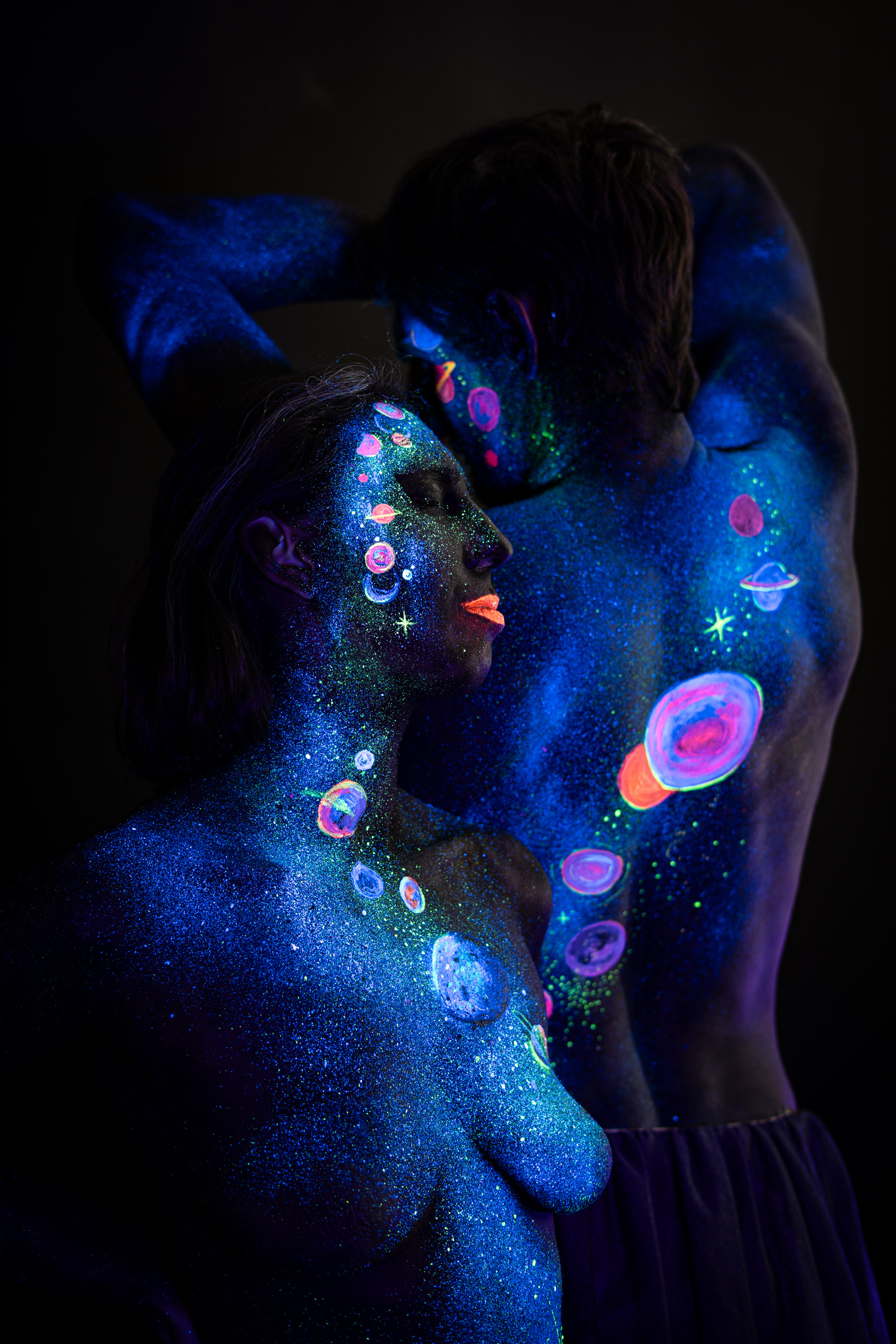 Two models, Alessia standing in the foreground in a three-quarter view facing the camera and Axell standing behind her with the back turned. A shared space themed design of glowing planets covers both bodies. Bodypaint by Elad Itzkin, Charlotte Bobineau, Sage Brewer. Photographed by Elad Itzkin Photography under UV/blacklight in his studio in Gesundbrunnen, Berlin.