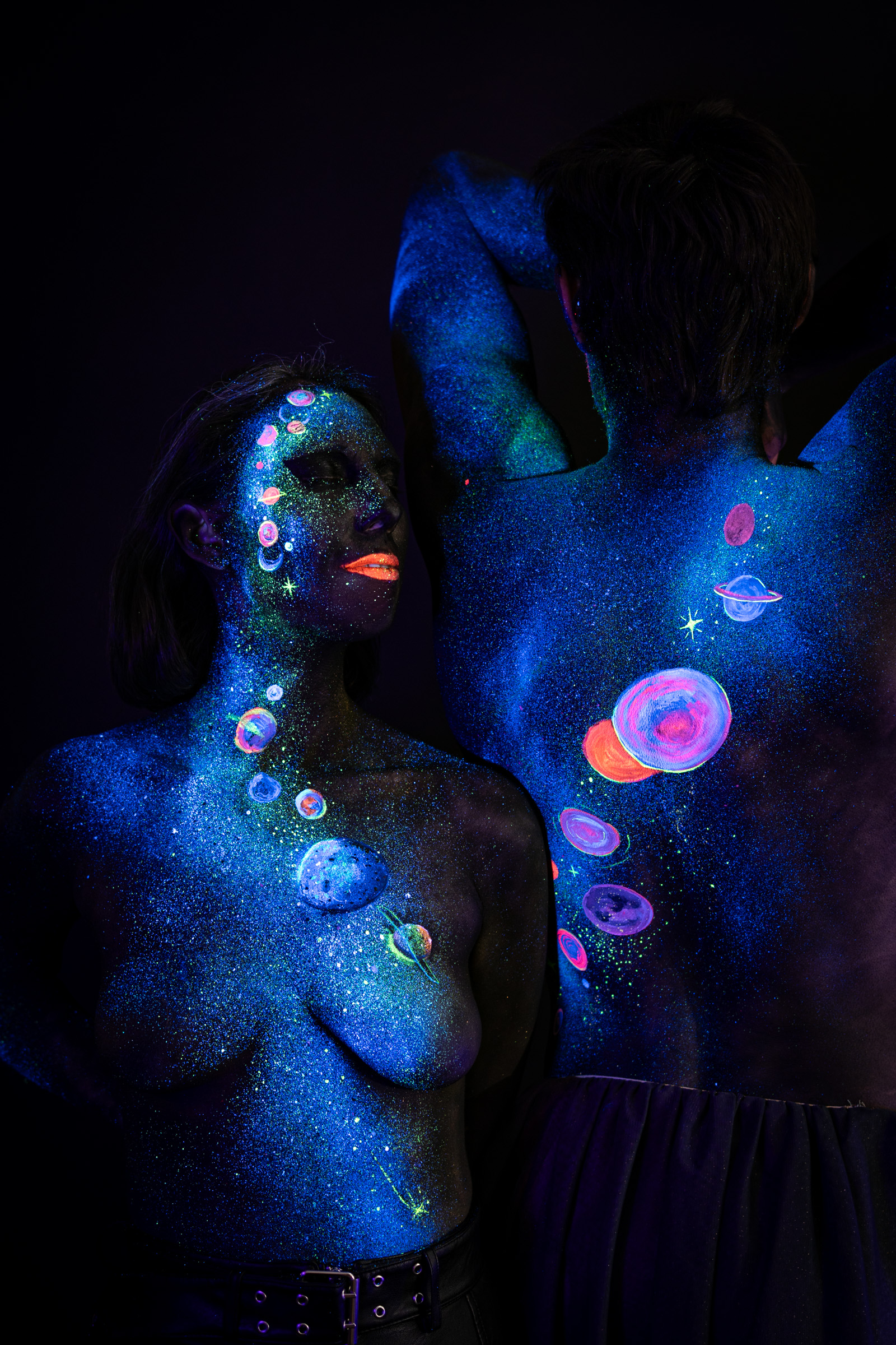 Two models, Alessia standing in front in a three-quarter pose and Axell standing behind her with the back to the camera, a variation of the same composition. Glowing planets in pink, orange and blue mark both bodies. Bodypaint by Elad Itzkin, Charlotte Bobineau, Sage Brewer. Photographed by Elad Itzkin Photography under UV/blacklight in his studio in Gesundbrunnen, Berlin.