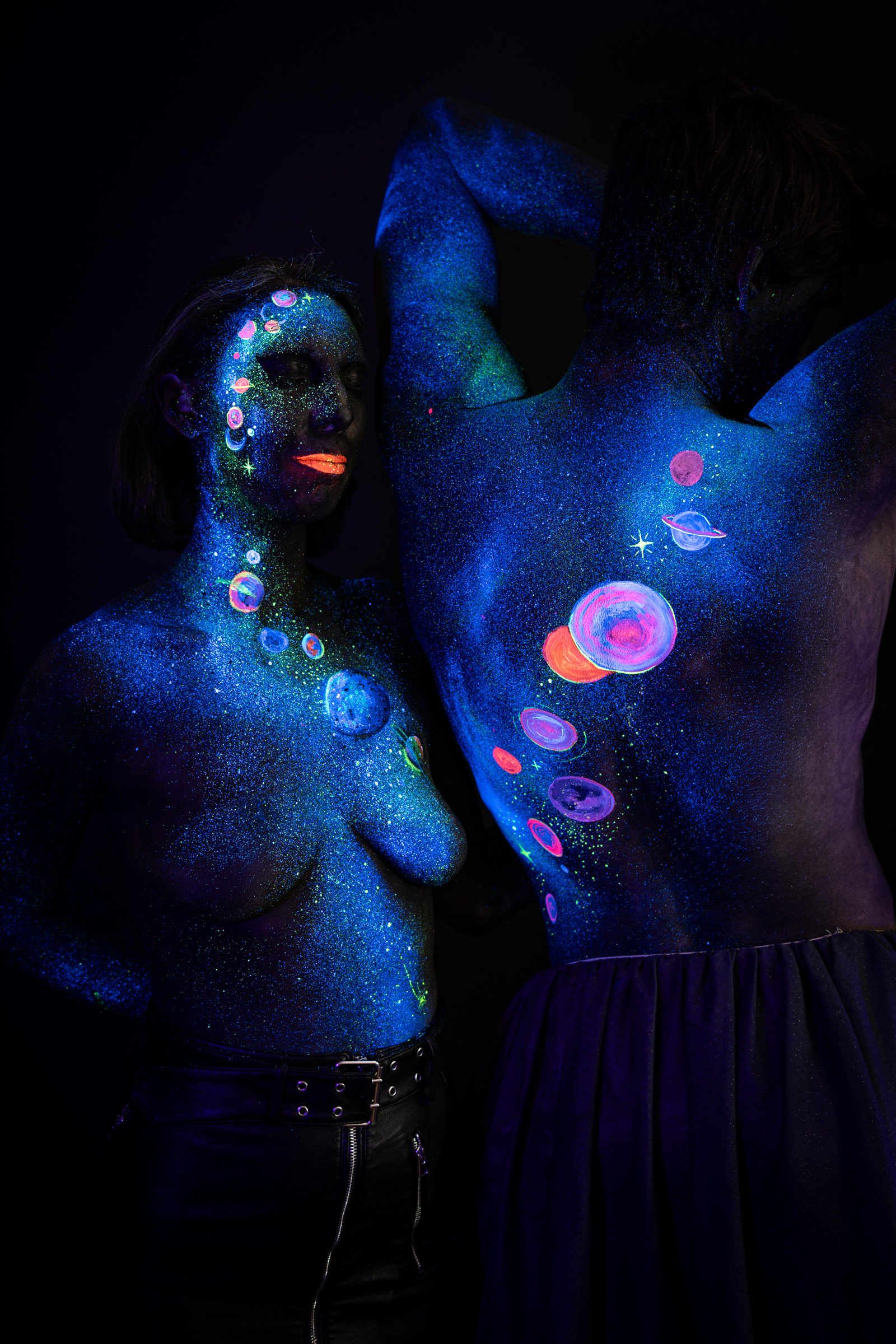 Alessia stands in three-quarter view in front of Axell, who stands behind her with his back turned to the camera. Both are painted in a blue-toned cosmic design with pink, orange and purple planets and stars over a glittering starfield. Bodypaint by Elad Itzkin, Charlotte Bobineau, Sage Brewer. Photographed by Elad Itzkin Photography under UV/blacklight in his studio in Gesundbrunnen, Berlin.