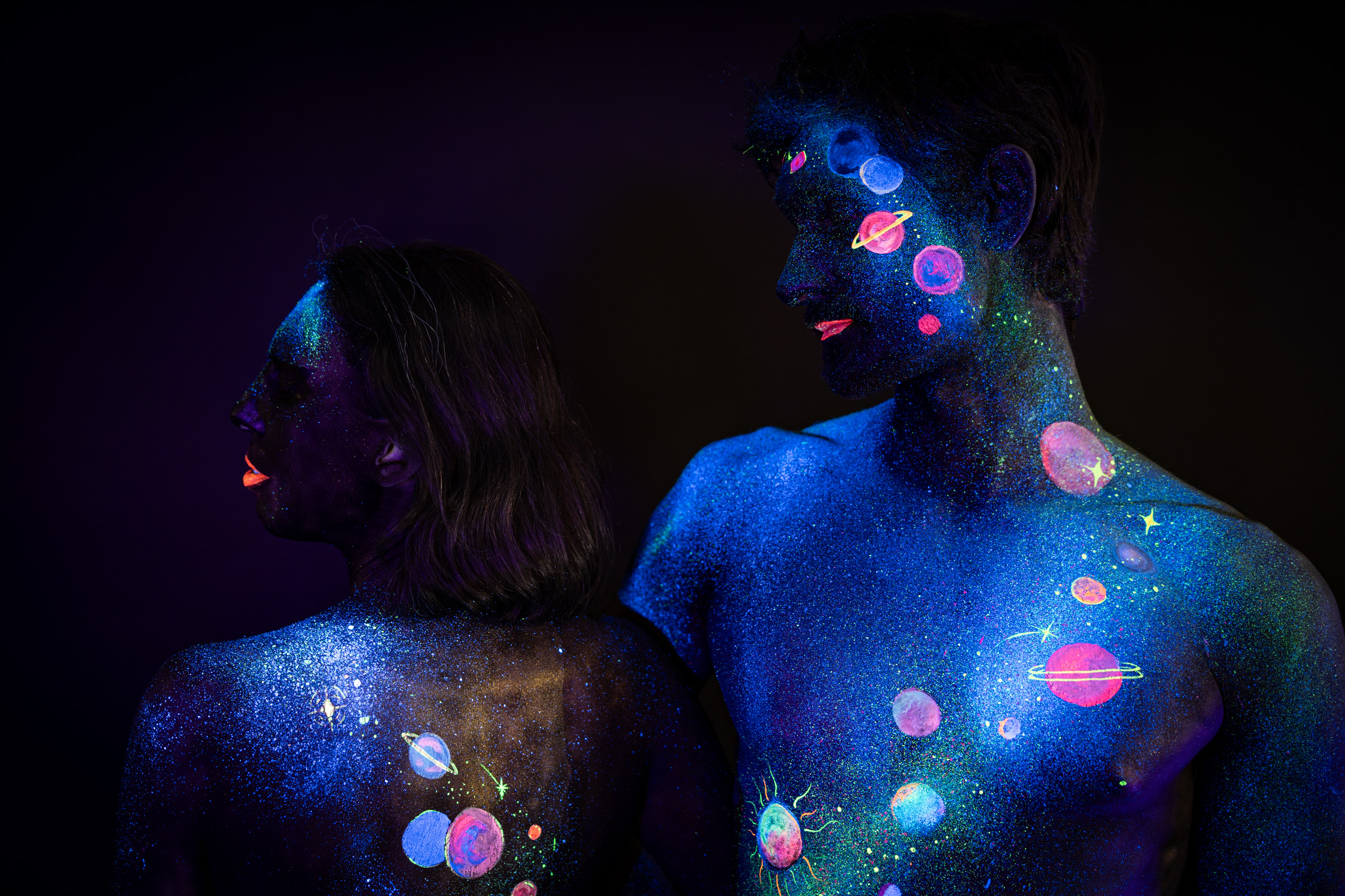 Alessia and Axell are photographed from behind, both in three-quarter turned poses with their faces angled to the side. Their bodypaint depicts a galaxy motif of glowing planets and stars in pink, orange and green against a blue glittered background. Bodypaint by Elad Itzkin, Charlotte Bobineau, Sage Brewer. Photographed by Elad Itzkin Photography under UV/blacklight in his studio in Gesundbrunnen, Berlin.