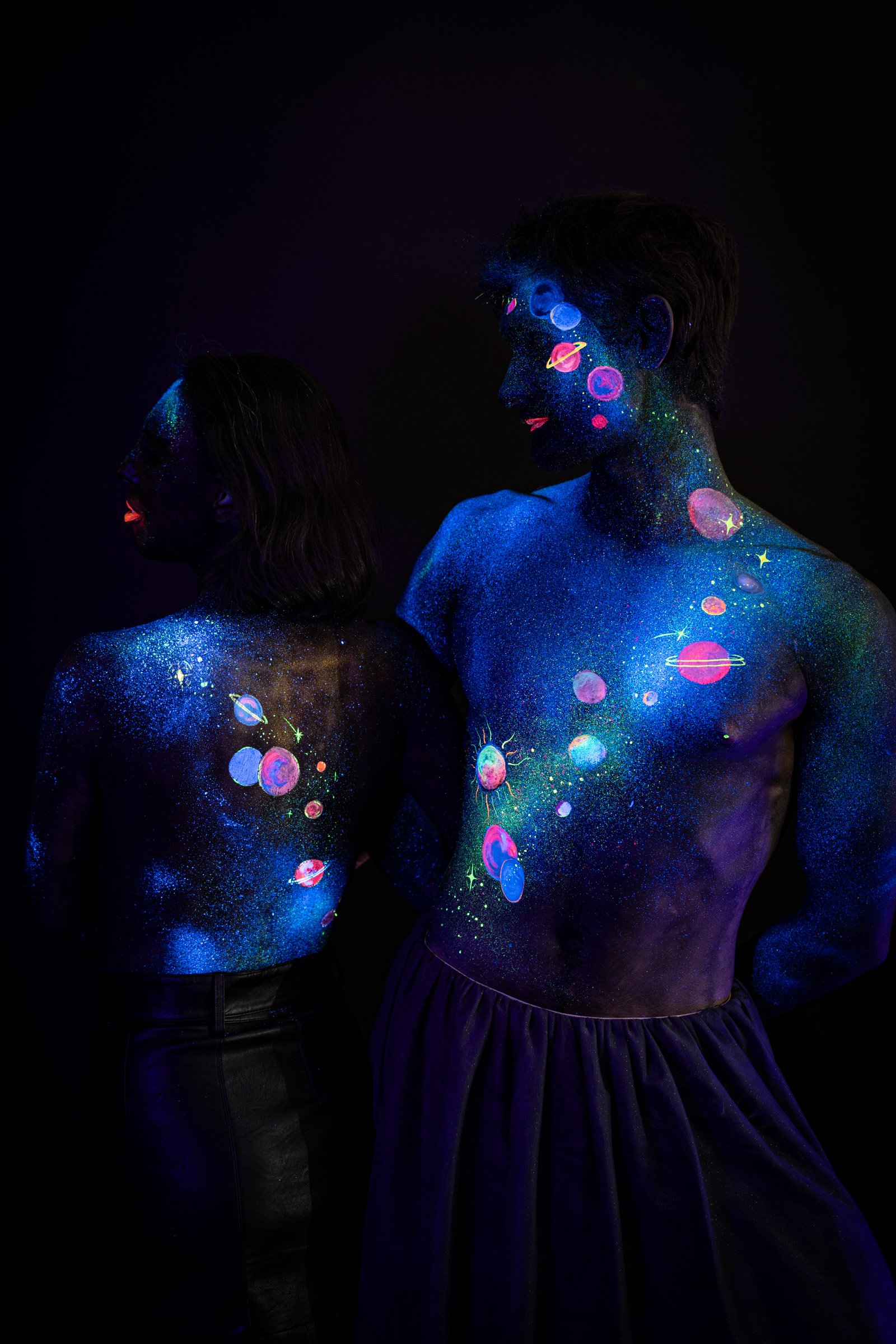 Axell stands in three-quarter view facing to the side, with Alessia standing close behind him, her back turned to the camera. Both bodies are painted with a space themed design of colorful planets and stars over a blue glitter base. Bodypaint by Elad Itzkin, Charlotte Bobineau, Sage Brewer. Photographed by Elad Itzkin Photography under UV/blacklight in his studio in Gesundbrunnen, Berlin.
