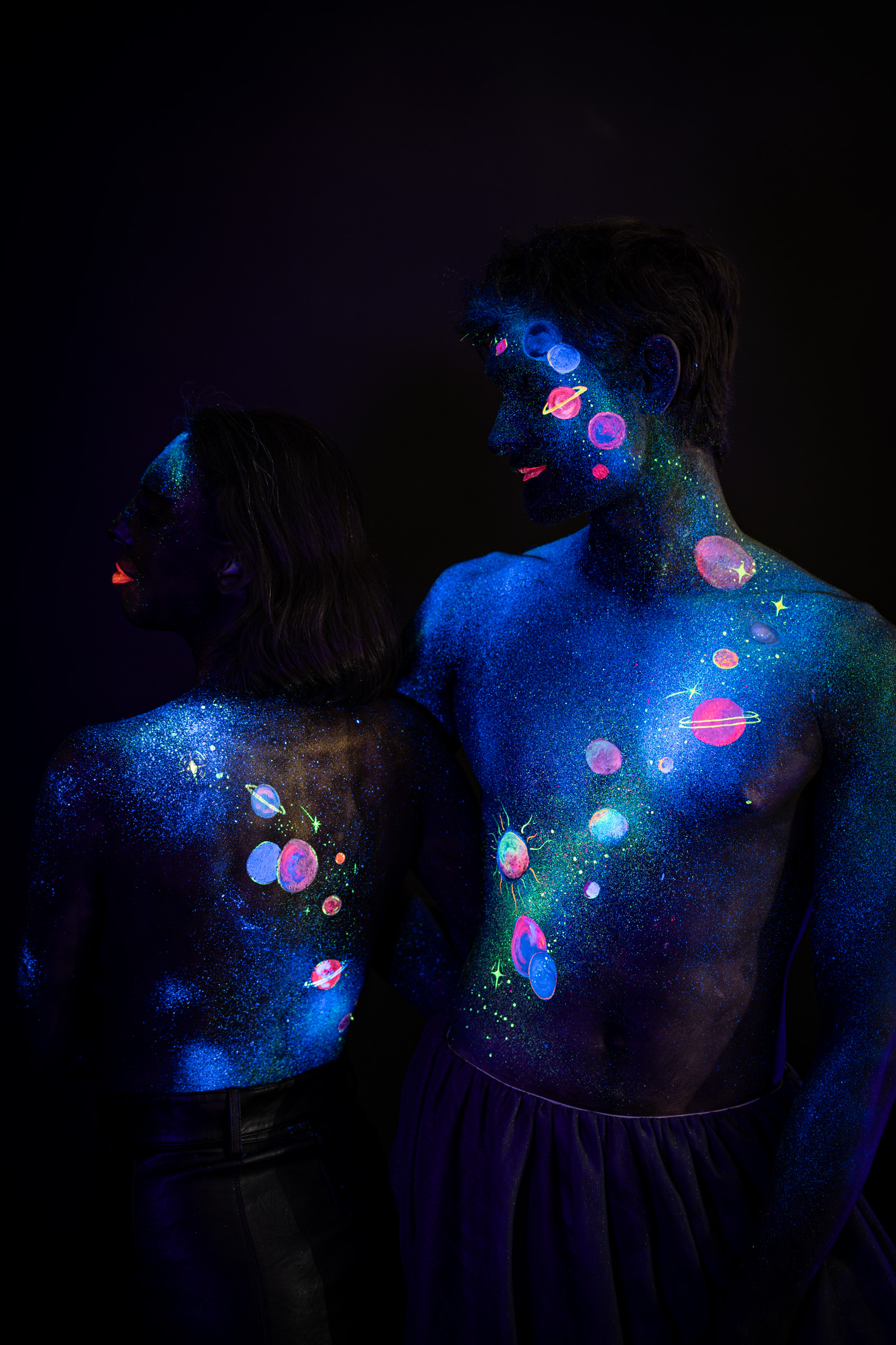 Alessia and Axell stand close together with their backs turned to the camera, their painted torsos and shoulders filling the frame. The design shows glowing pink, orange and purple planets and stars scattered across a blue glitter base. Bodypaint by Elad Itzkin, Charlotte Bobineau, Sage Brewer. Photographed by Elad Itzkin Photography under UV/blacklight in his studio in Gesundbrunnen, Berlin.