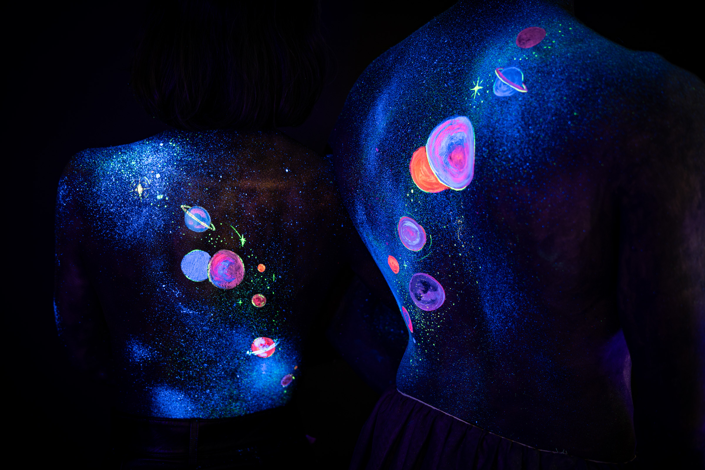 This close view shows Alessia and Axell standing with their backs to the camera, upper torsos filling the frame. Their bodypaint is a cosmic design of orange, pink and purple planets and rings set against a starry blue glitter background. Bodypaint by Elad Itzkin, Charlotte Bobineau, Sage Brewer. Photographed by Elad Itzkin Photography under UV/blacklight in his studio in Gesundbrunnen, Berlin.