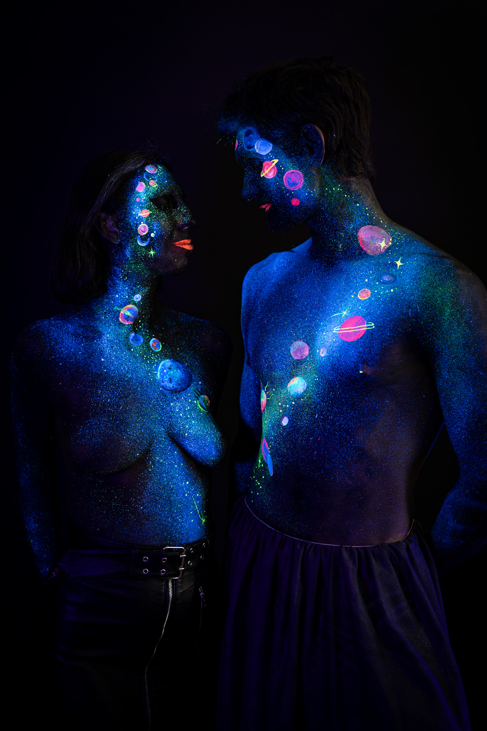 Alessia and Axell stand facing each other, their painted faces close together. The bodypaint is a cosmic pattern of luminous planets, rings and scattered stars in pink, orange and purple over a blue glitter base. Bodypaint by Elad Itzkin, Charlotte Bobineau, Sage Brewer. Photographed by Elad Itzkin Photography under UV/blacklight in his studio in Gesundbrunnen, Berlin.