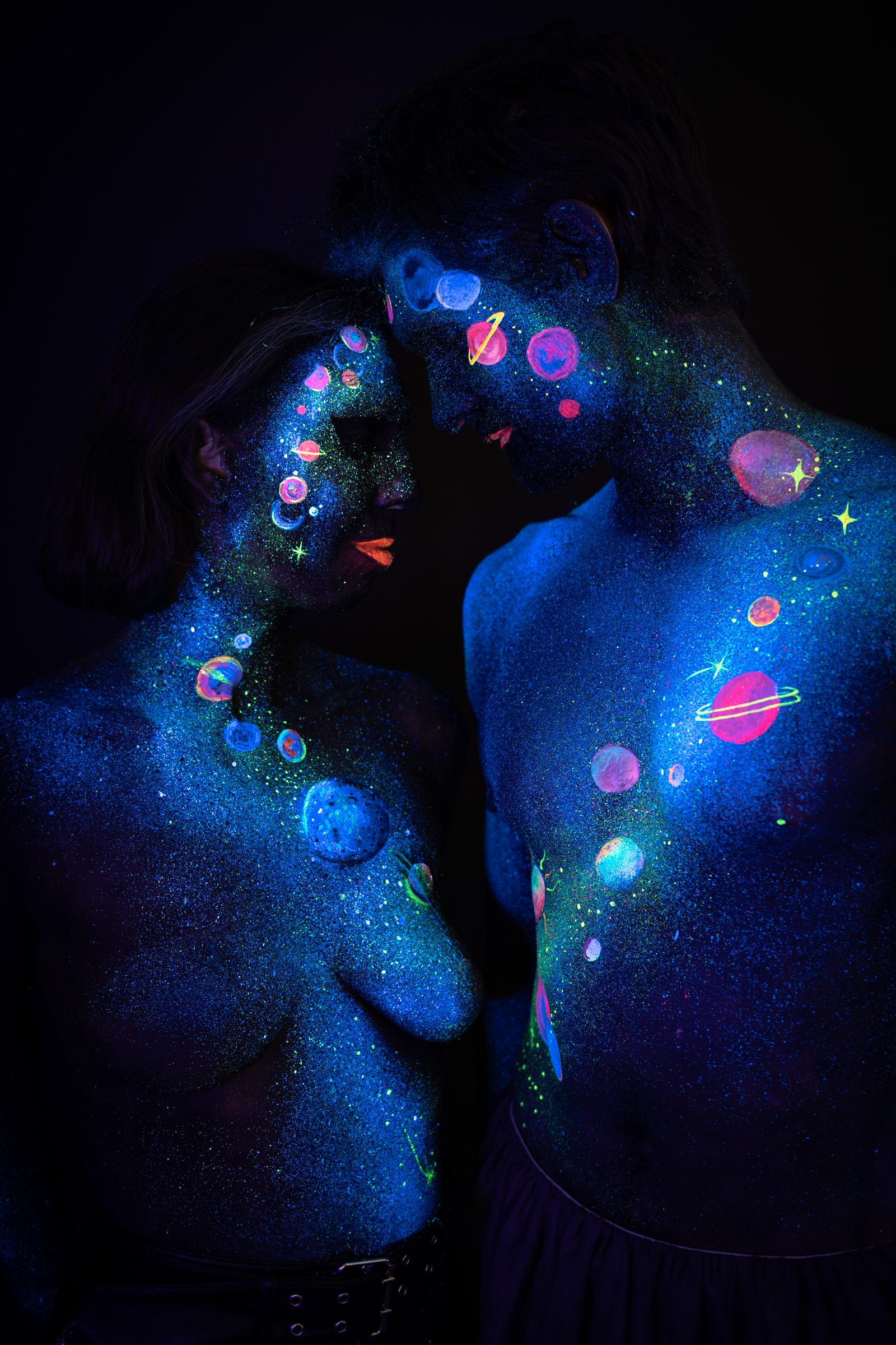 In this wider shot, Alessia and Axell stand facing each other with their foreheads close together. The cosmic bodypaint design includes glowing planets, a ringed sphere and scattered stars in pink, orange and green over blue glitter. Bodypaint by Elad Itzkin, Charlotte Bobineau, Sage Brewer. Photographed by Elad Itzkin Photography under UV/blacklight in his studio in Gesundbrunnen, Berlin.