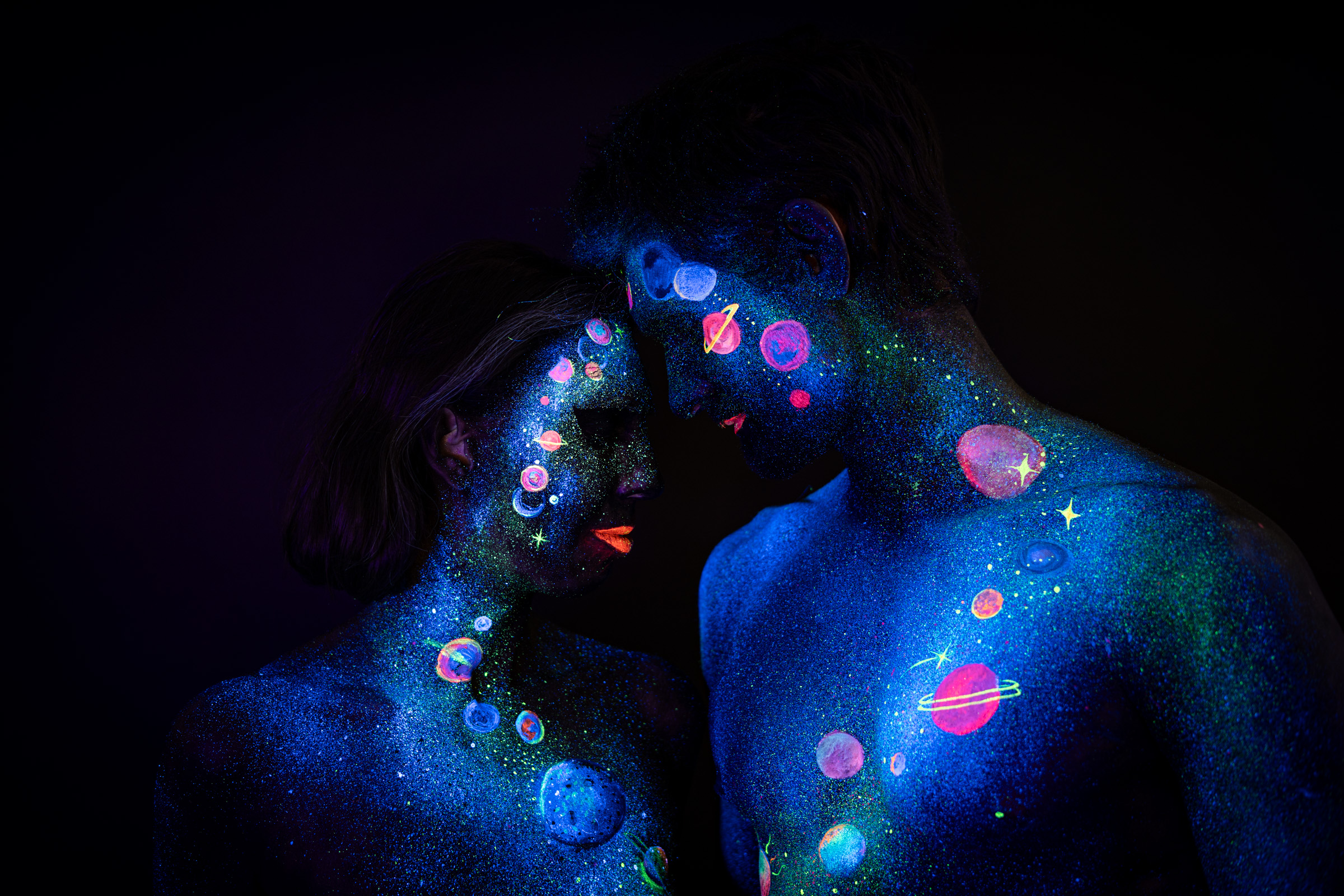 Alessia and Axell stand facing each other, foreheads almost touching, with their painted torsos visible below. The bodypaint depicts a galaxy scene of orange, pink and purple planets and stars over a blue glitter background. Bodypaint by Elad Itzkin, Charlotte Bobineau, Sage Brewer. Photographed by Elad Itzkin Photography under UV/blacklight in his studio in Gesundbrunnen, Berlin.