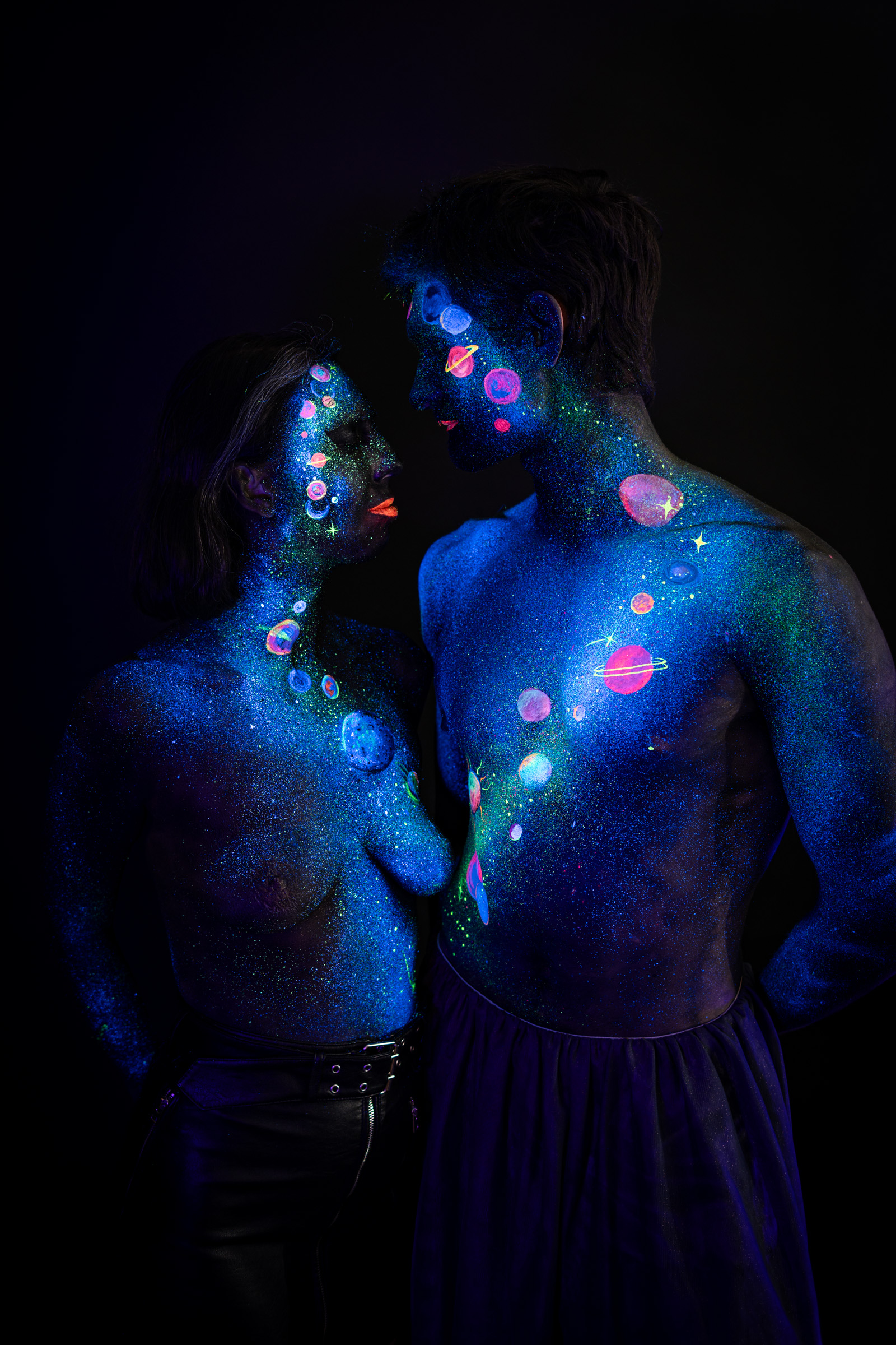 This close up shows Alessia and Axell standing face to face with their foreheads touching. Their bodypaint is a cosmic design of glowing planets, rings and stars in pink, orange and green over a deep blue glitter base. Bodypaint by Elad Itzkin, Charlotte Bobineau, Sage Brewer. Photographed by Elad Itzkin Photography under UV/blacklight in his studio in Gesundbrunnen, Berlin.