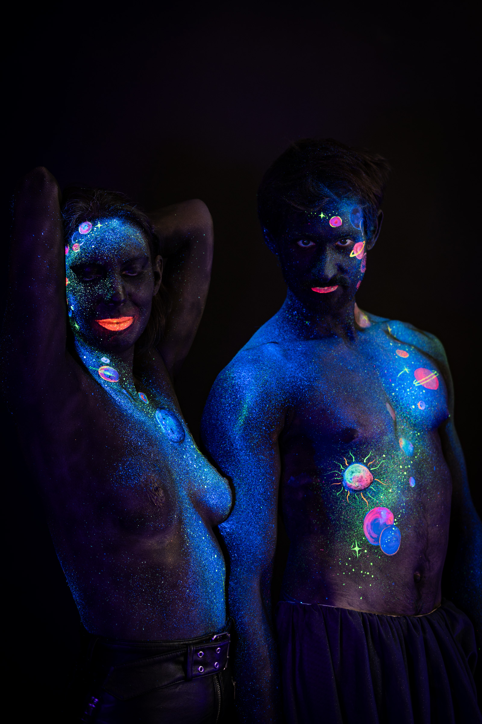 Alessia stands facing toward the camera in a three-quarter pose, with Axell standing beside her also facing forward. The bodypaint on both models is a cosmic design of glowing orange, pink and purple planets and stars over blue glitter. Bodypaint by Elad Itzkin, Charlotte Bobineau, Sage Brewer. Photographed by Elad Itzkin Photography under UV/blacklight in his studio in Gesundbrunnen, Berlin.