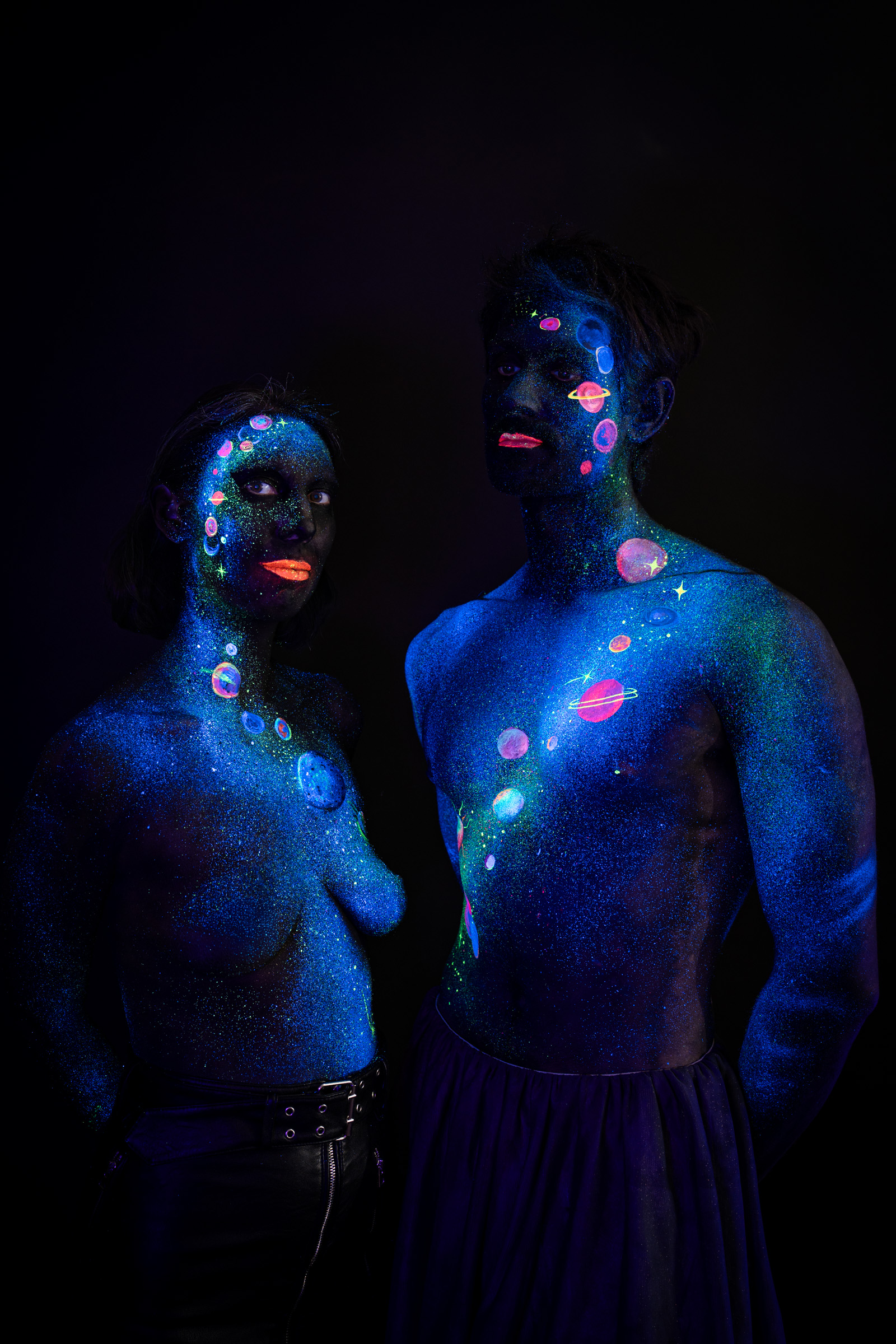 Alessia stands looking toward Axell, who faces forward toward the camera, both positioned close together. Their bodypaint depicts a cosmic scene of orange, pink and green planets and stars over a blue glitter background. Bodypaint by Elad Itzkin, Charlotte Bobineau, Sage Brewer. Photographed by Elad Itzkin Photography under UV/blacklight in his studio in Gesundbrunnen, Berlin.